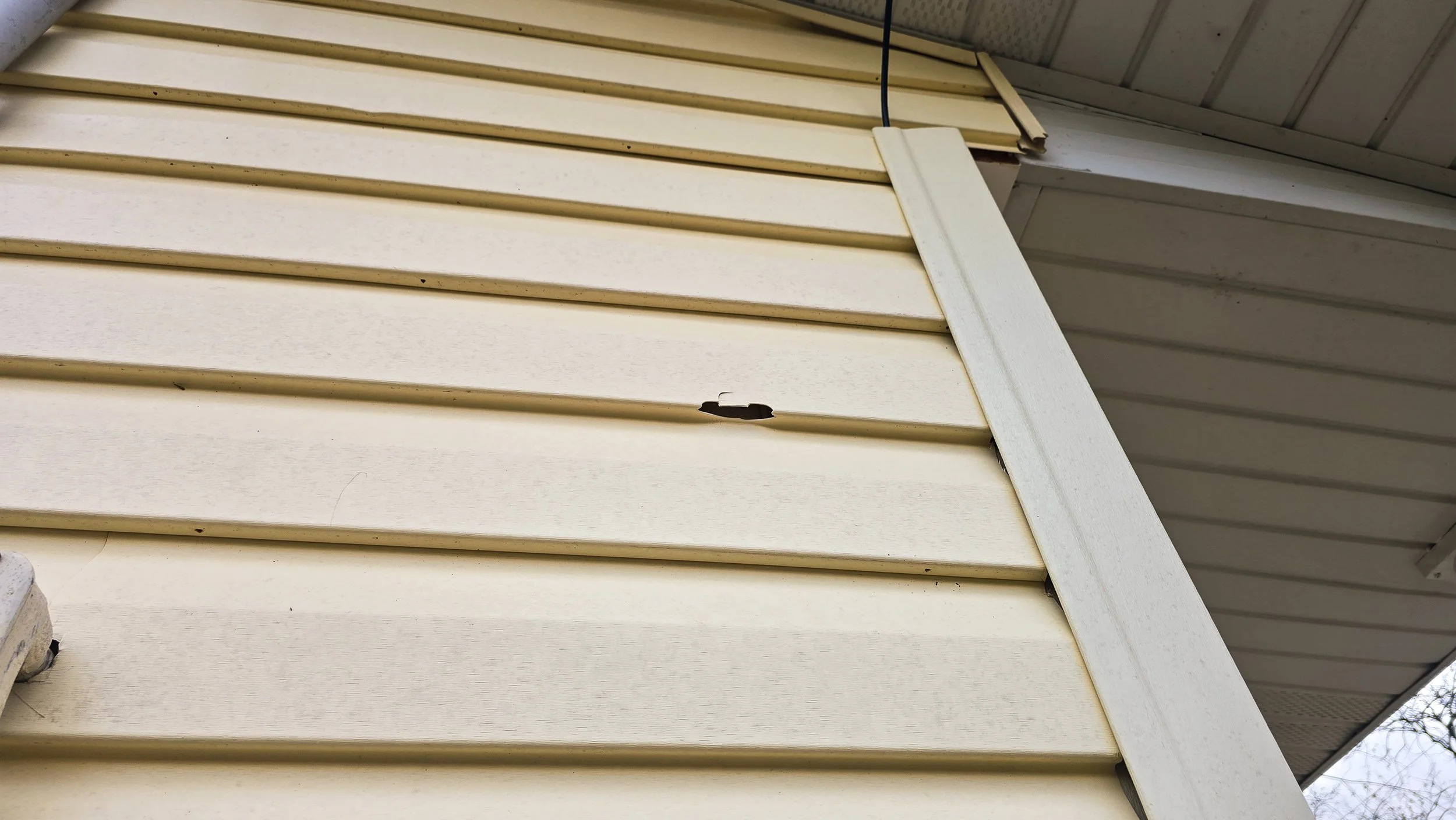 Close-up of beige vinyl siding with a small hole or damage near the middle, and white soffit extending from the upper right corner.