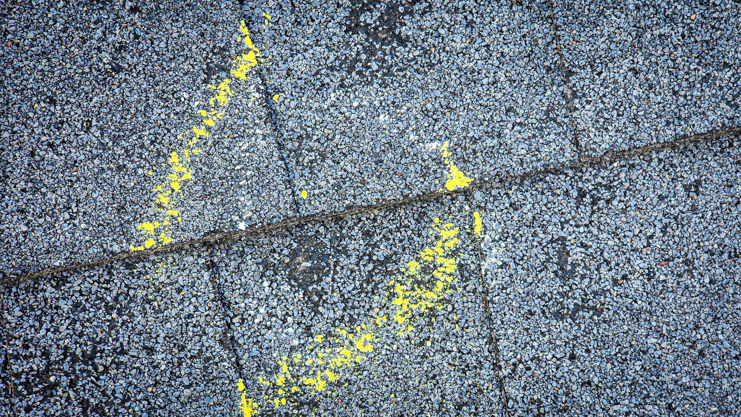 Hail impact. Hail damage. Hail to the roof. Hail damage to shingles. Gray asphalt pavement with yellow spray-painted arrow markings.