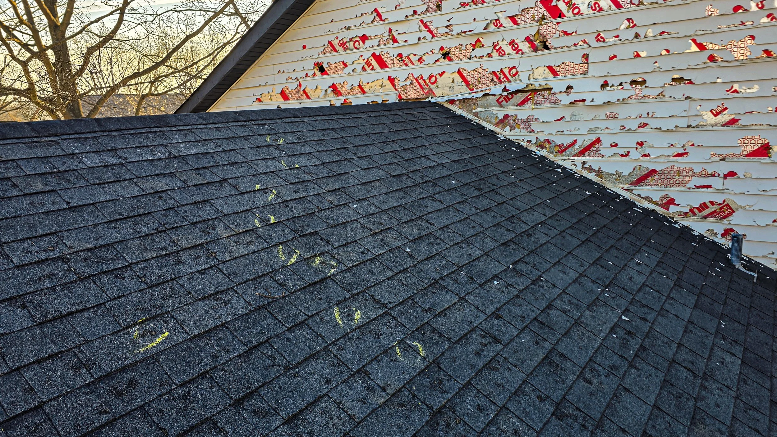 Partial view of a roof with dark asphalt shingles and a neighboring house being renovated, with its siding partially removed, revealing insulation and structural elements.