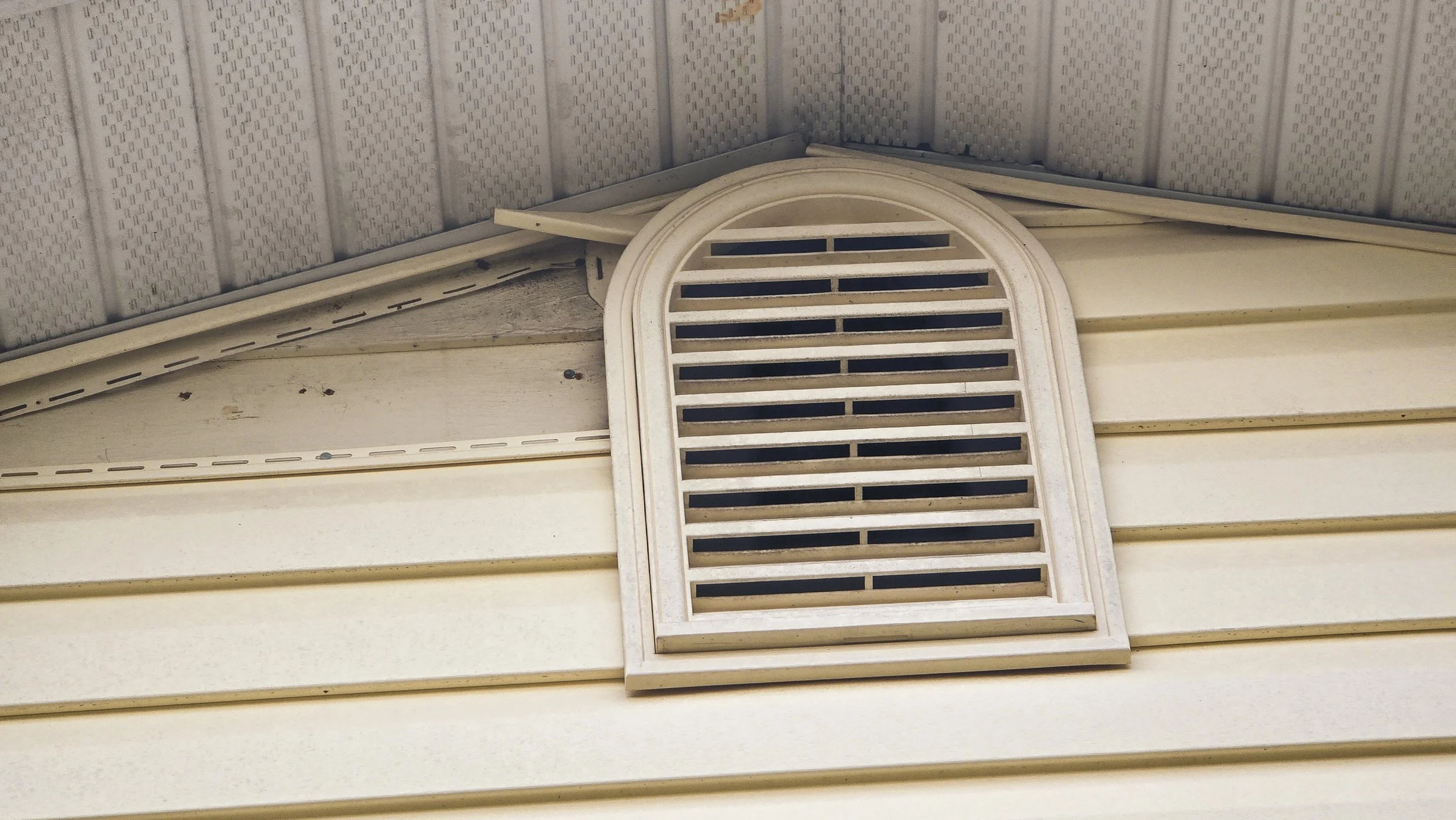 An outdoor wall vent with a decorative arched top, mounted on a beige horizontal siding wall with a textured ceiling above.