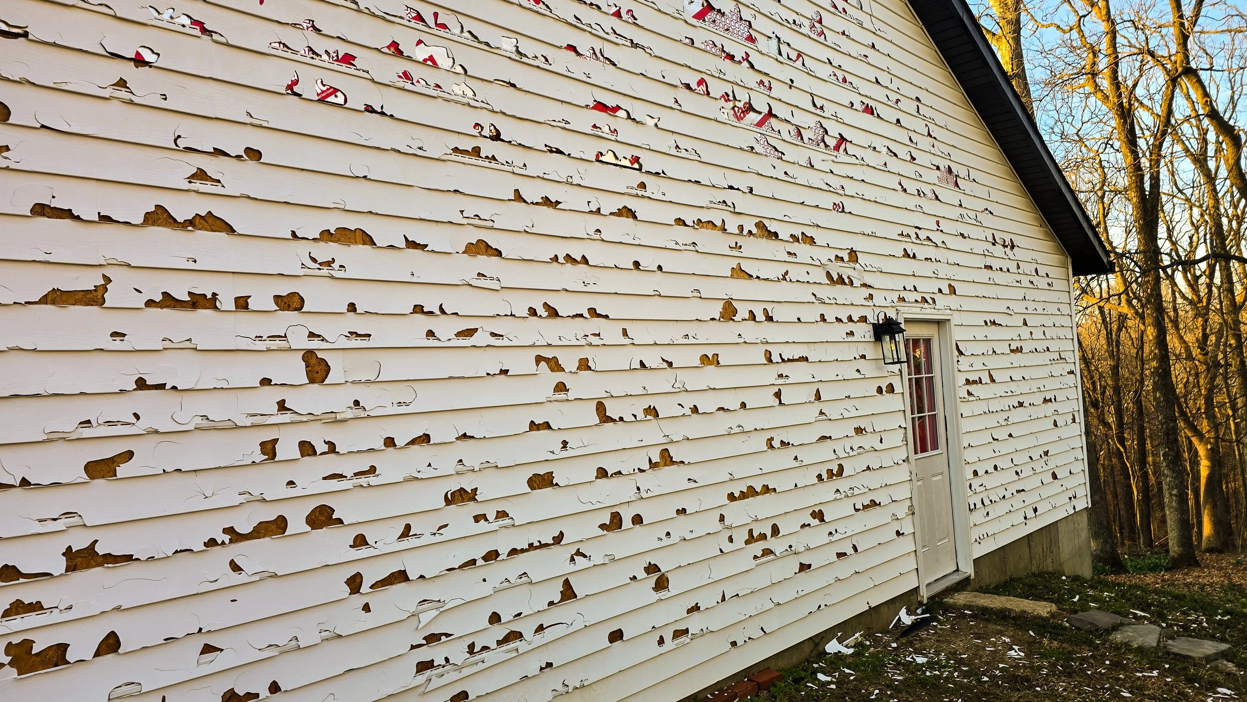 Siding of a house with peeling and damaged white paint, exposing the wood underneath.