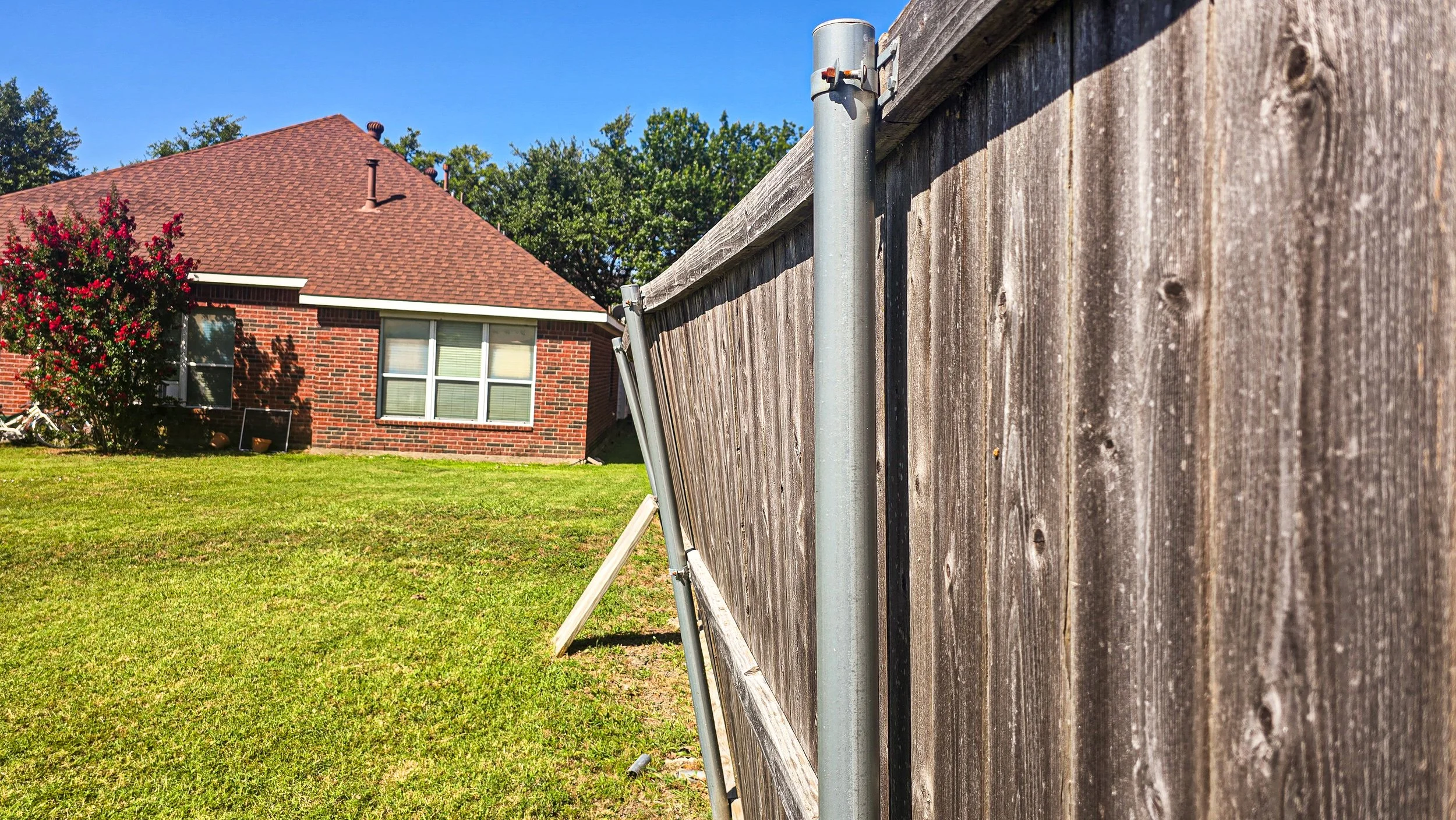 A wooden fence on the right side of the image, with a leaning metal support pole. In the background, a brick house with a red shingle roof, windows, and a bush with pink flowers. The sky is clear and blue, with green trees in the distance.