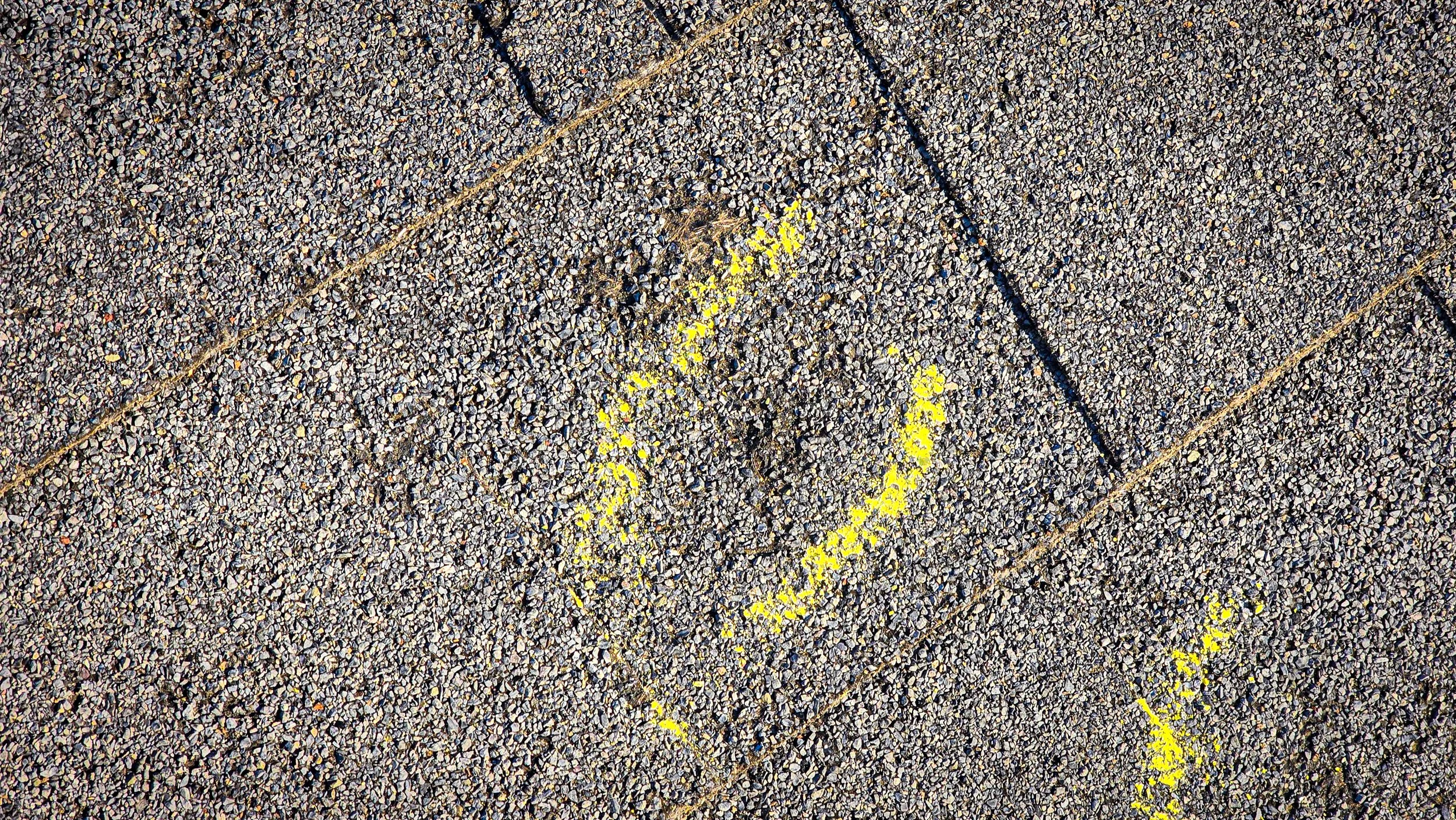 Hail impact. Hail damage. Hail to the roof. Hail damage to shingles. Yellow paint markings on gray asphalt shoulder.