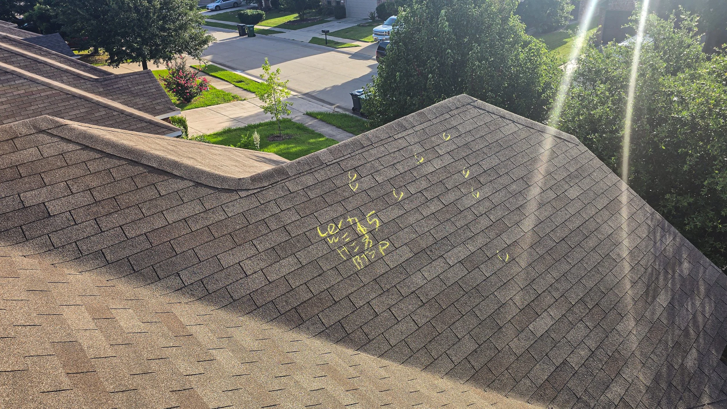 View of a residential neighborhood with a brown shingle roof, trees, lawns, sidewalks, and parked cars, taken from the roof amid bright sunlight.