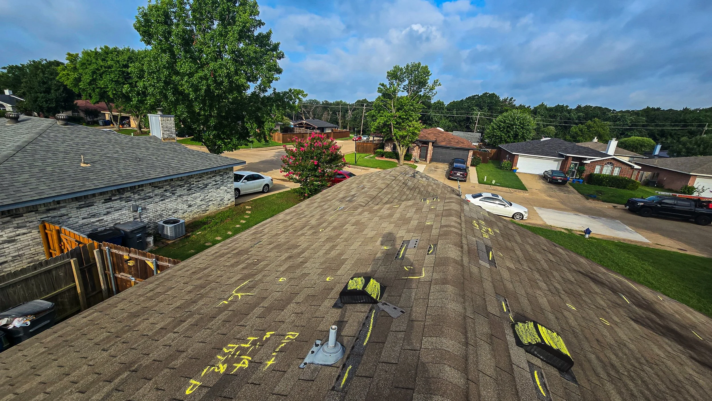 View from a rooftop showing a neighborhood with houses, trees, parked cars, and a partly cloudy sky.
