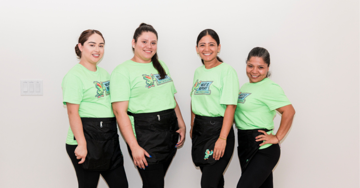 Four women standing in a row against a plain white wall, smiling, wearing matching neon green shirts with a logo and black aprons.