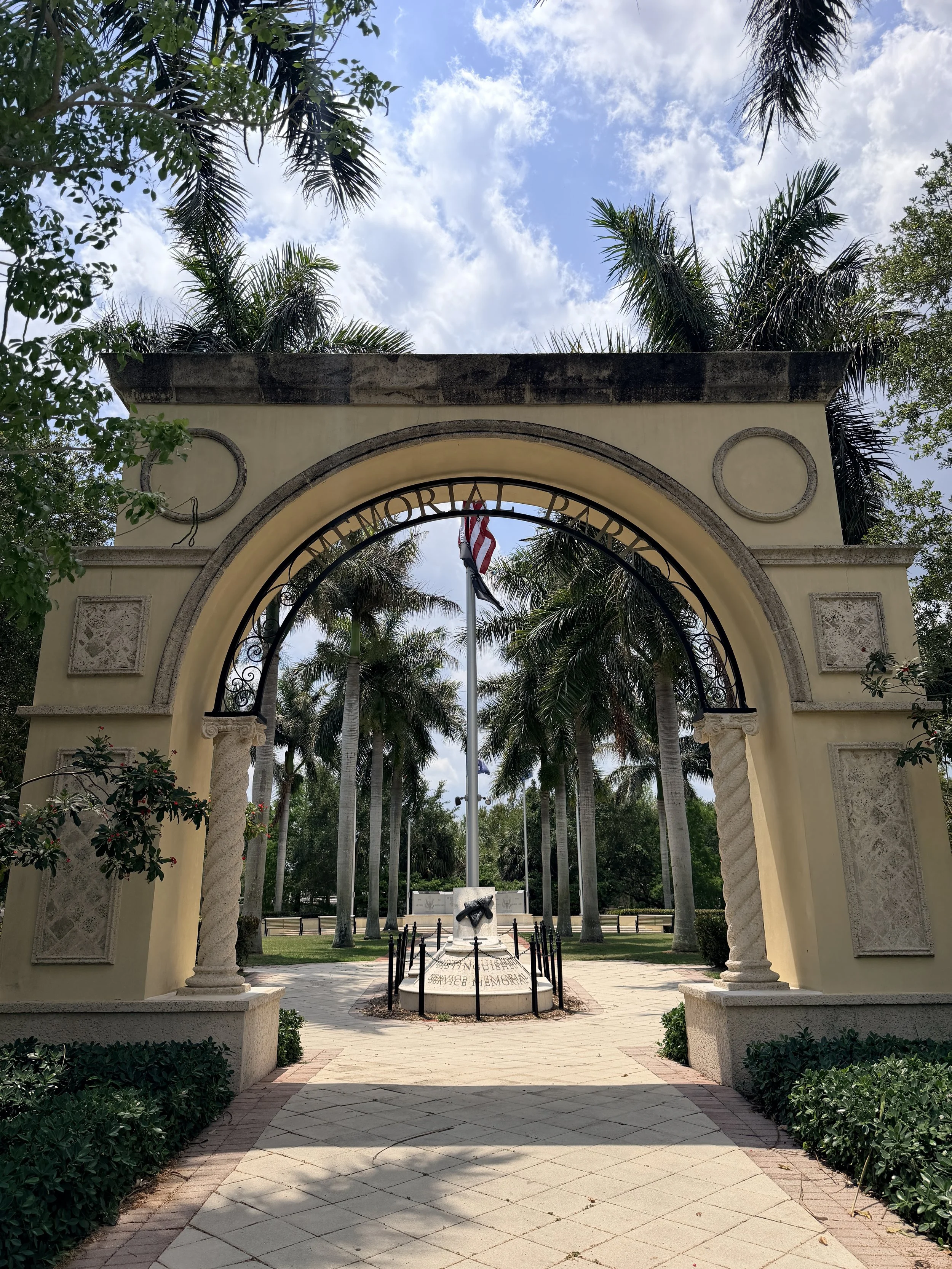 An archway with the words Memorial Park, flanked by columns with circular decorative elements, leading to a park with palm trees, a flagpole, and a sculpture in a memorial area.
