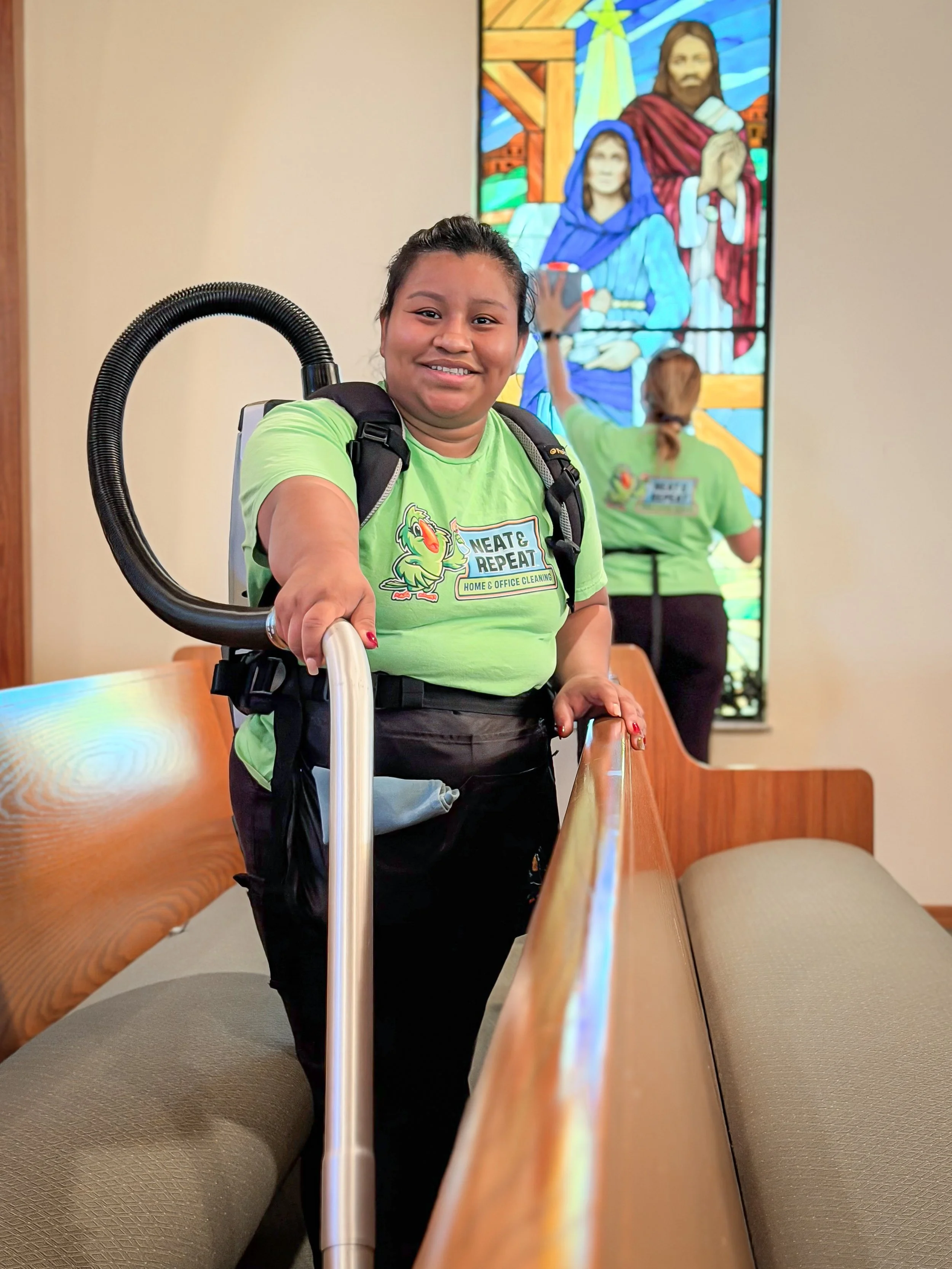 A woman with a backpack and a cleaning uniform smiling and holding onto a handrail inside a church. There is a stained-glass window with religious images in the background, and another person in a similar uniform cleaning the window.