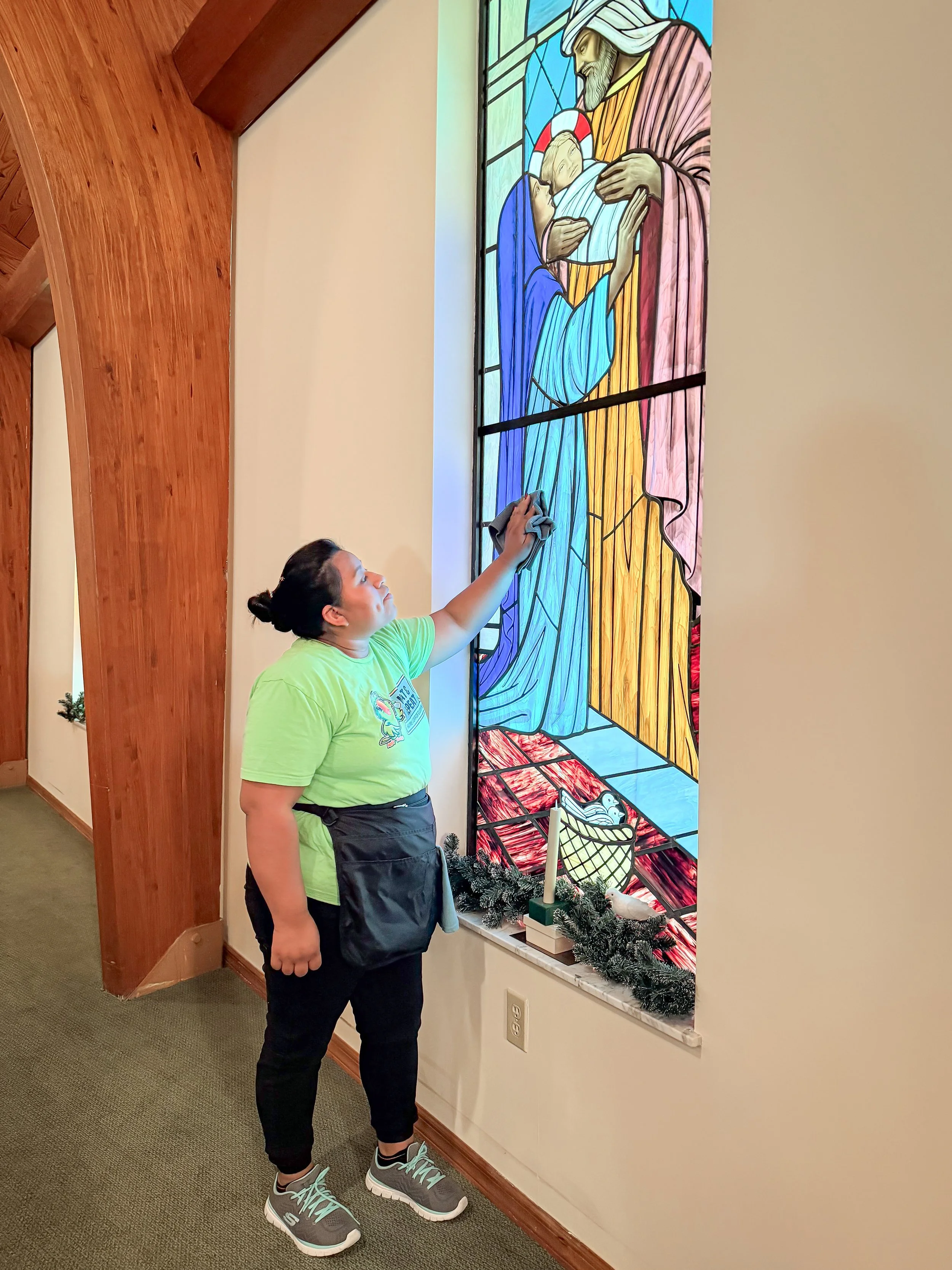 A woman cleaning a colorful stained glass window depicting the Nativity scene with Mary, Joseph, baby Jesus, a manger, and other details, in a church or chapel setting.