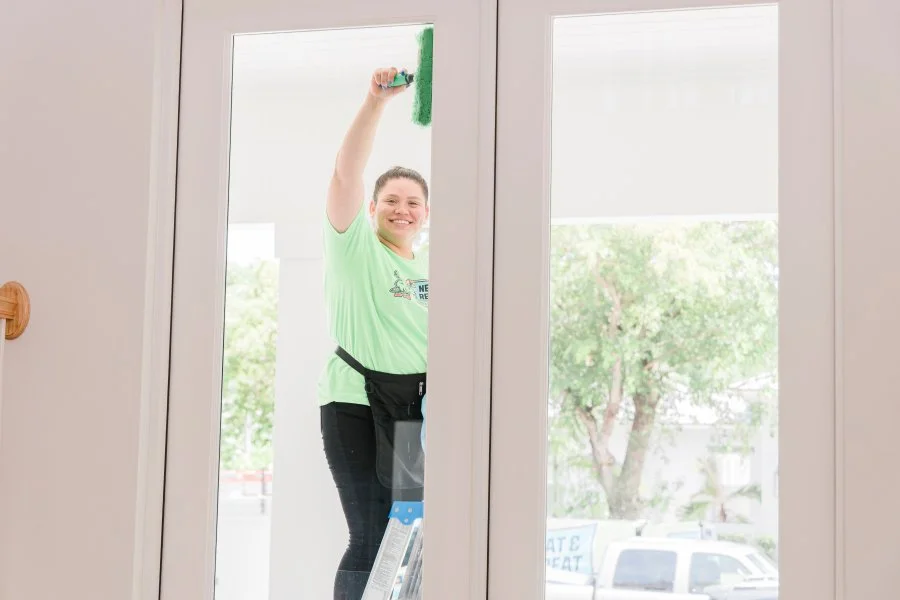 A woman smiling and painting the glass door with green paint from the outside.