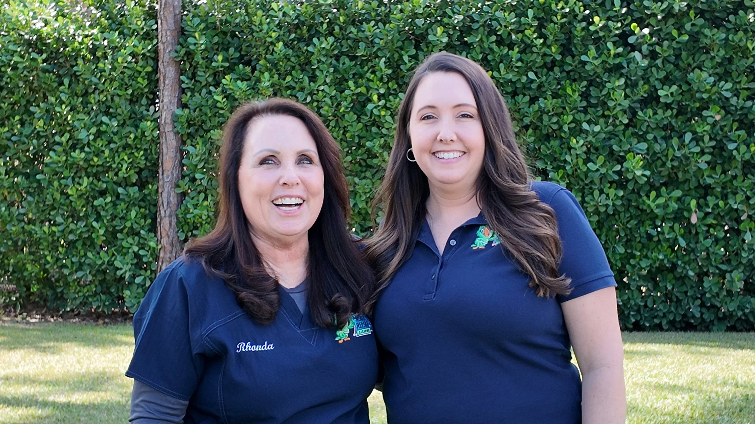 Two women standing outdoors in front of green bushes, both wearing navy blue shirts with embroidered logos, smiling and posing for the photo.