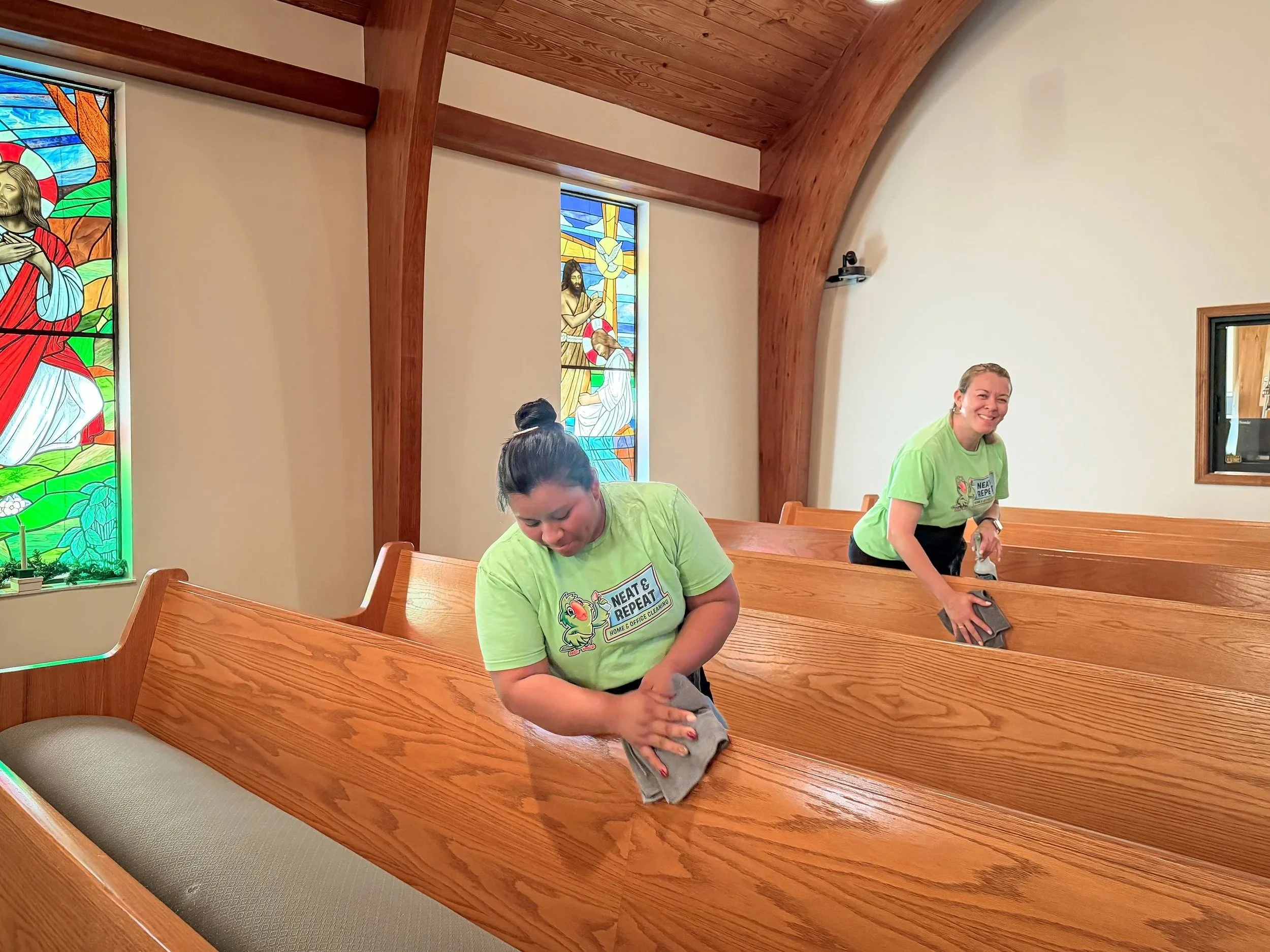 Two women wearing green shirts cleaning wooden pews inside a church with stained glass windows depicting religious figures.