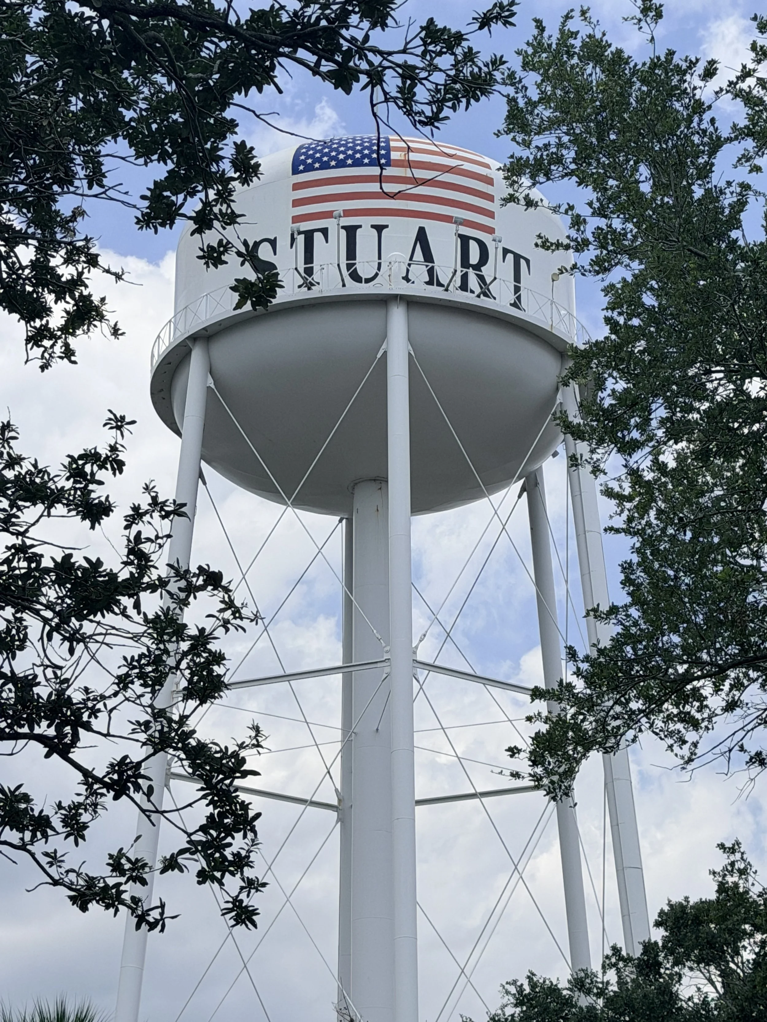A white water tower with the word "STUART" and an American flag painted on it, surrounded by trees and partly cloudy sky.