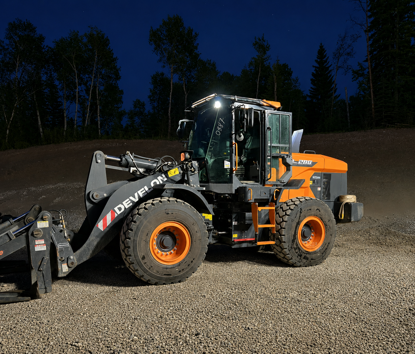 A large orange and black wheel loader with headlights on, positioned on a gravel surface at night, with a forested area and a dark blue sky in the background.