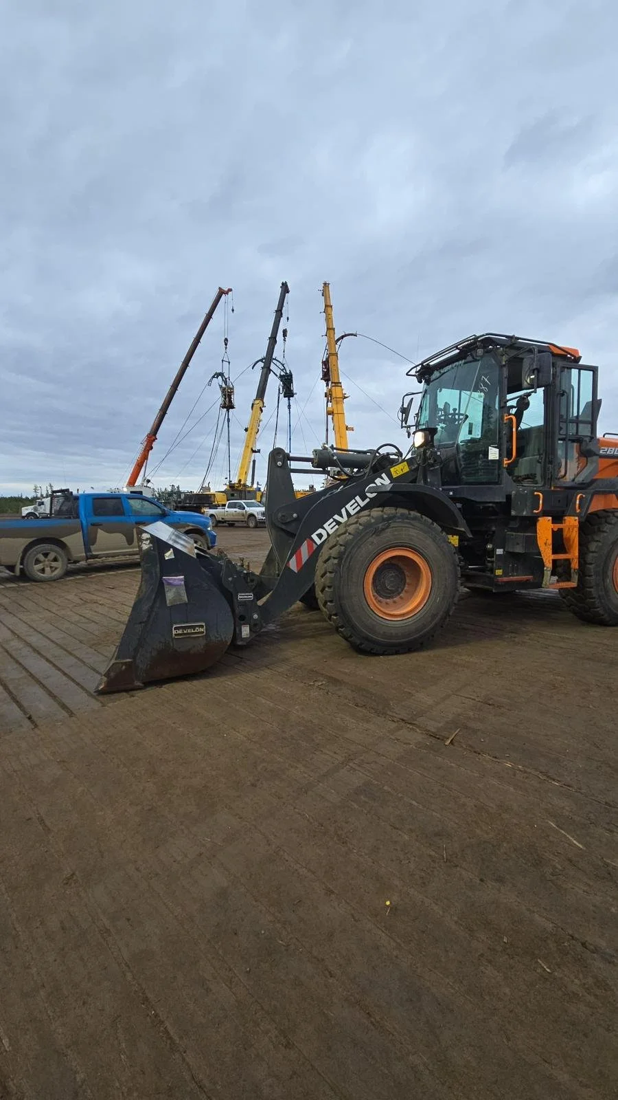 A large black and orange wheel loader with a front bucket attachment parked on a dirt surface at a construction site. Several cranes and trucks are visible in the background under a cloudy sky.