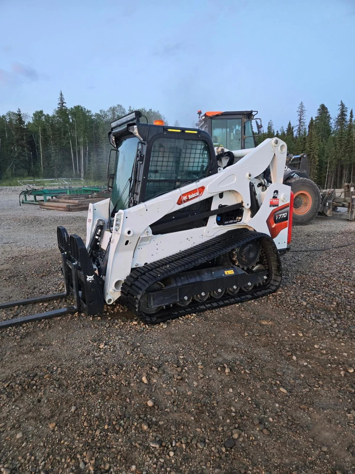 A Bobcat T770 compact track loader parked on gravel ground at a construction site with another larger piece of machinery in the background and a forest of trees beyond.