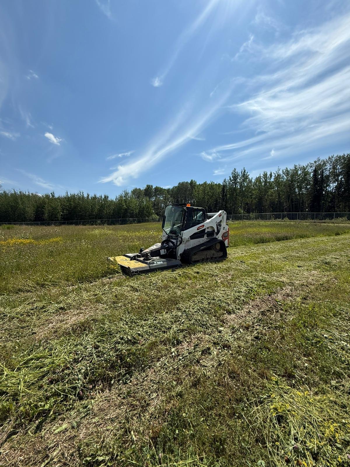 Small tracked mower working in a grassy field beneath a clear blue sky with sporadic clouds.