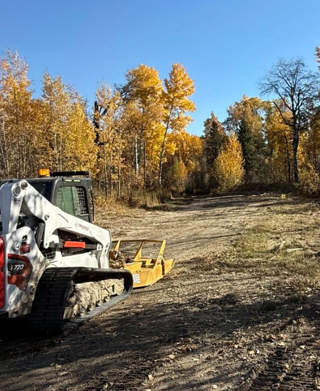 A construction vehicle parked on a dirt road surrounded by autumn-colored trees with a clear blue sky overhead.