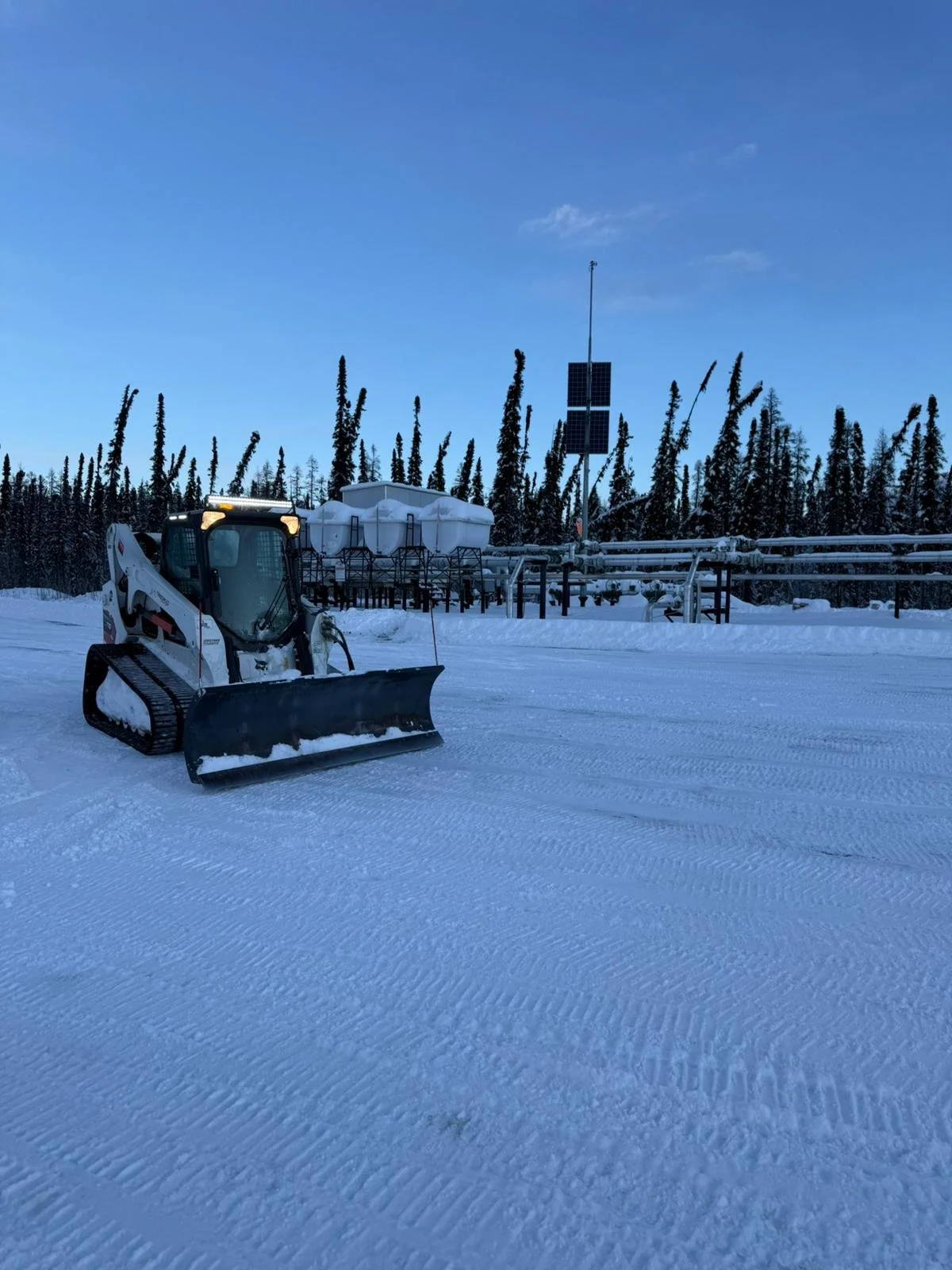 Snow-covered landscape with a small tracked snow removal vehicle and some equipment, trees, and a solar power system in the background under a blue sky.