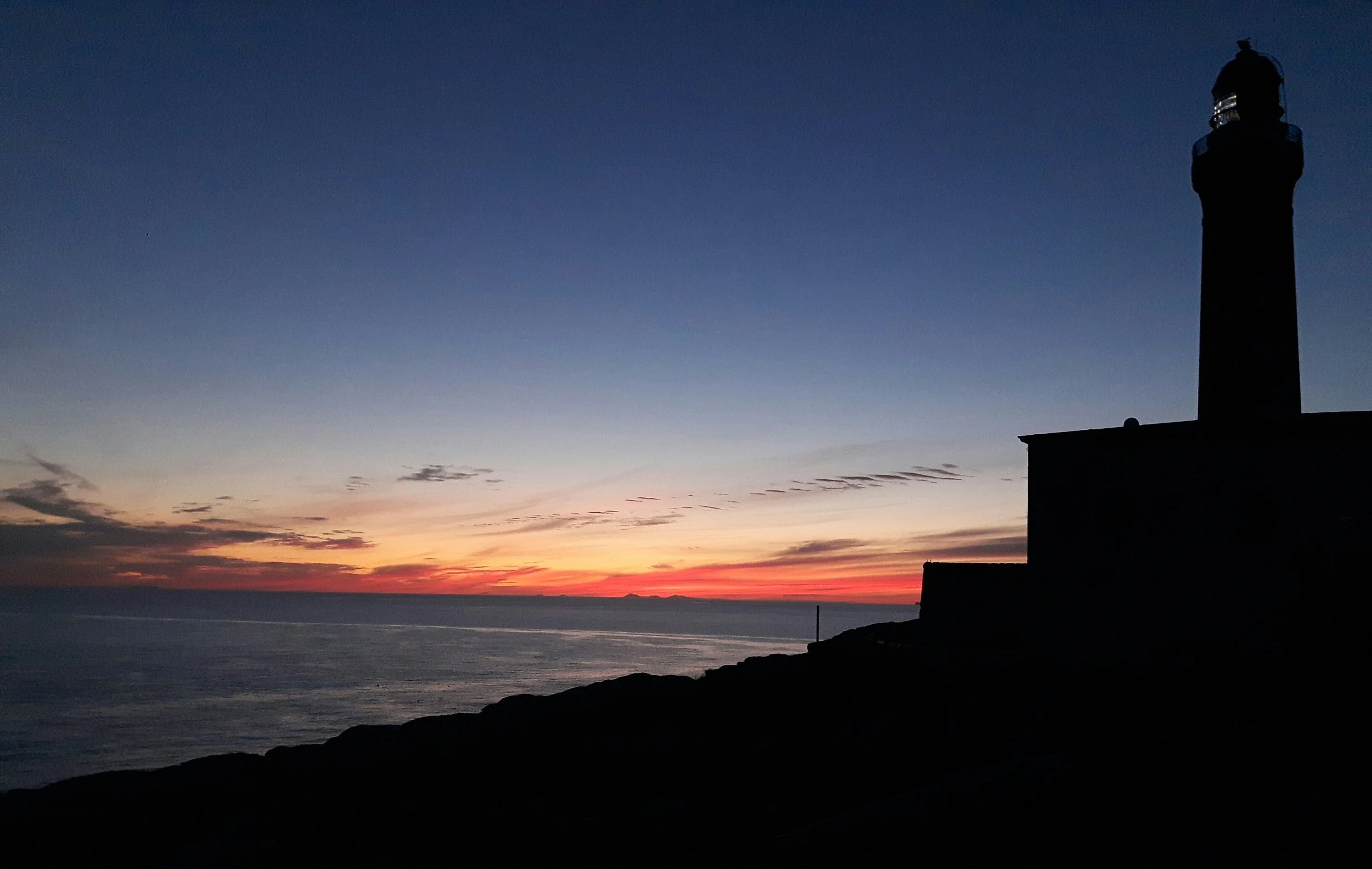 Silhouette of a lighthouse on a rocky shore at sunset or sunrise, with a colorful sky and calm ocean in the background.