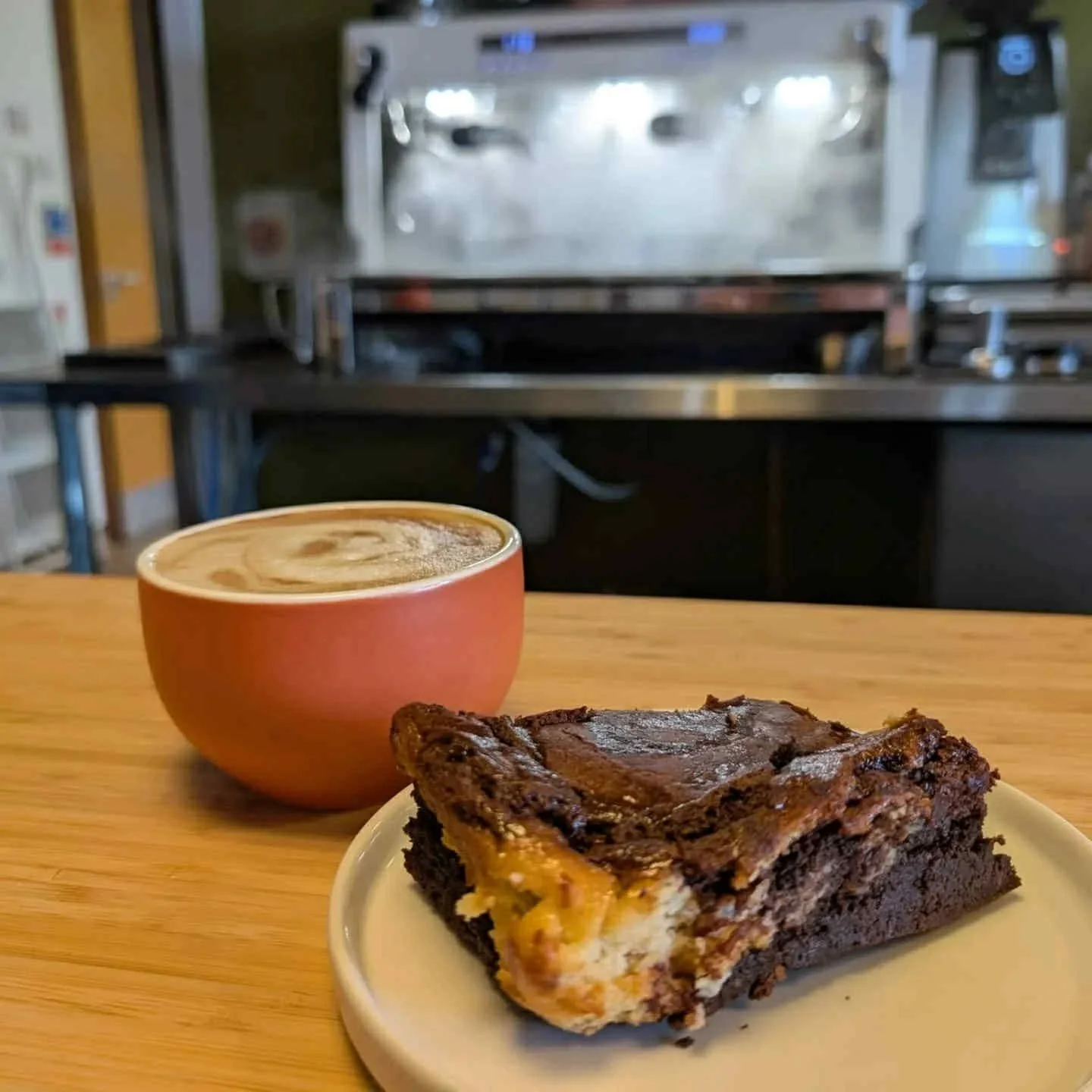 A cup of coffee and a chocolate cheesecake with yellow filling on a white plate, placed on a wooden table in front of a commercial kitchen oven.