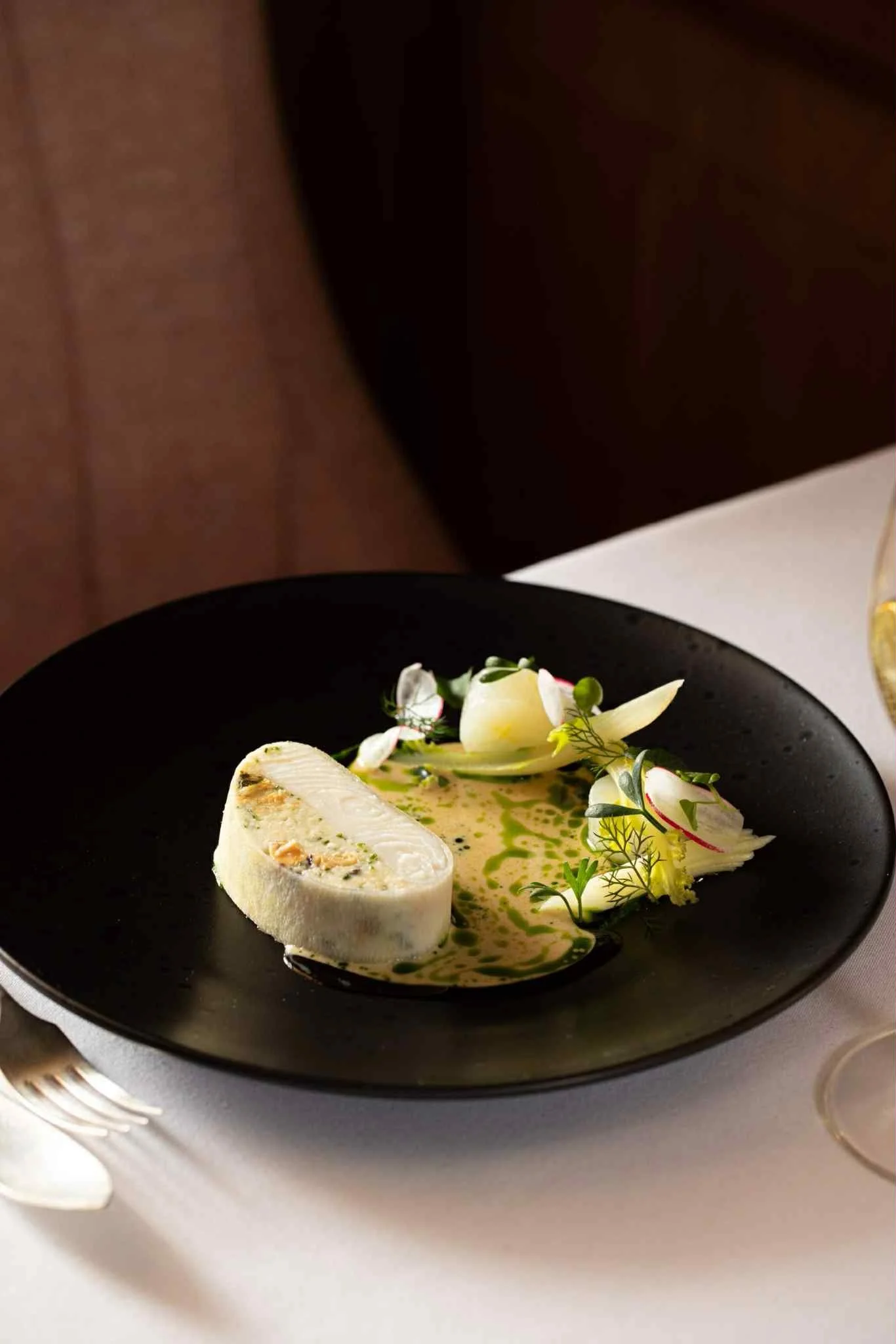 A gourmet dish featuring a sliced piece of creamy cheese on a black plate, garnished with edible herbs and flowers, served with a sauce.