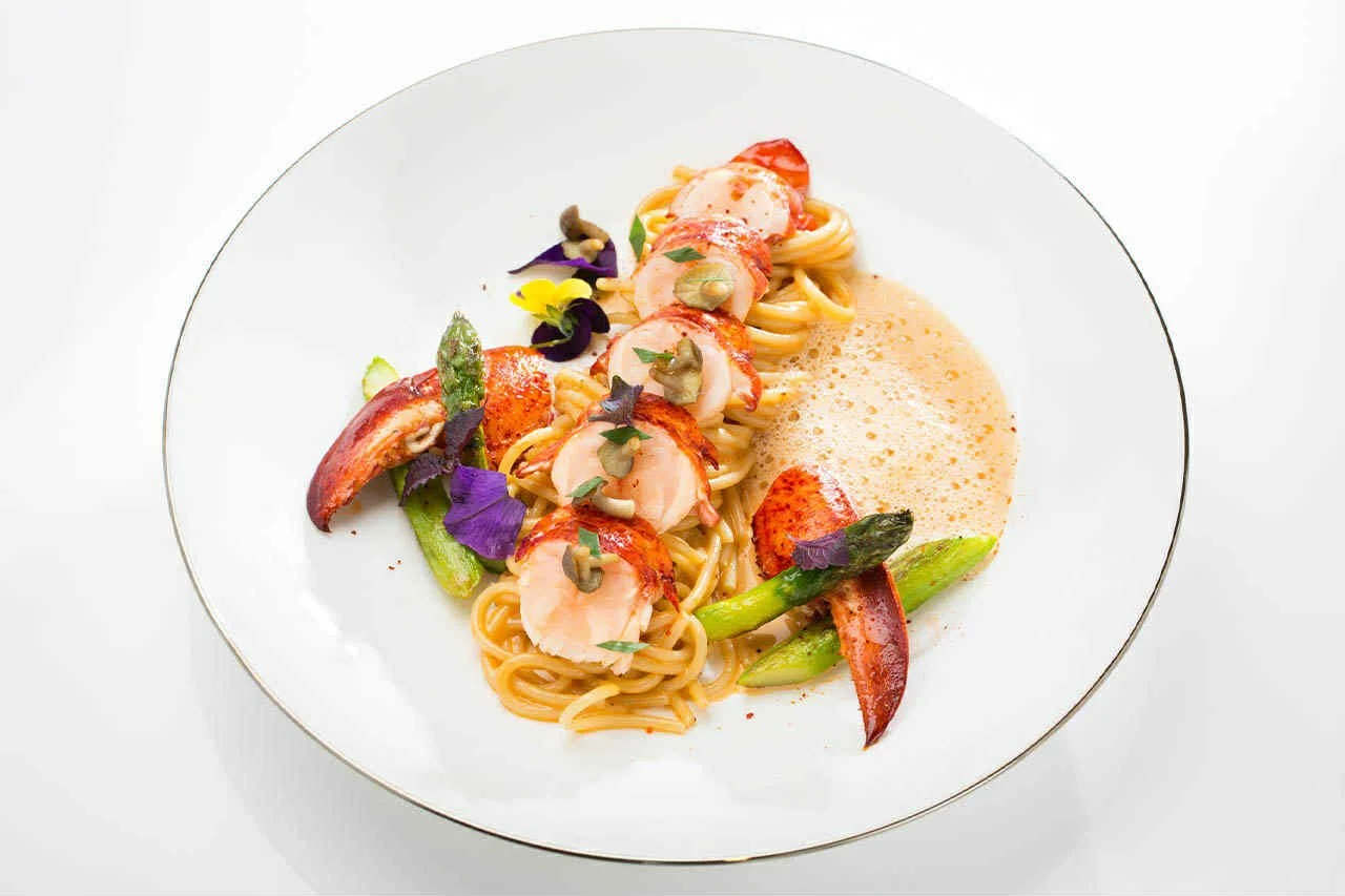 Plate of gourmet pasta with seared scallops, green asparagus, cherry tomatoes, edible flowers, and creamy sauce.