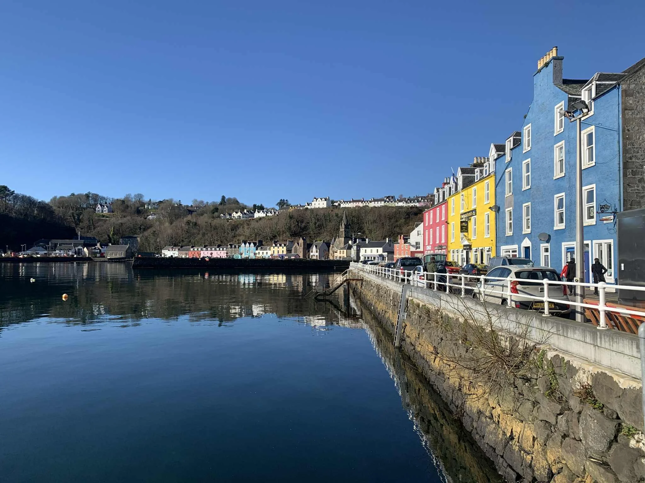 Colorful row of buildings in blue, yellow, and pink along a waterfront, with cars parked and pedestrians walking on the sidewalk, reflecting in the calm water under a clear blue sky.