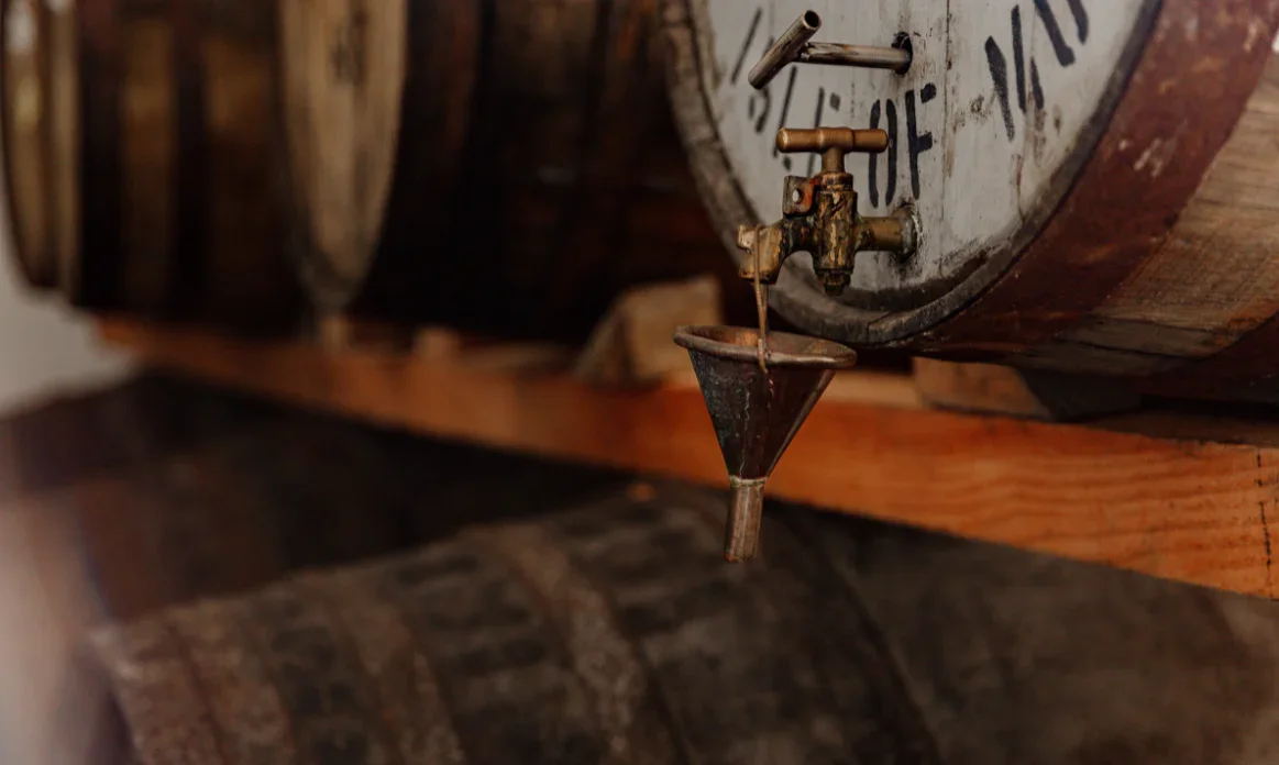 Close-up of an antique wooden clock with a visible clock face, rusty spigot, and a funnel hanging from it, all mounted on a wooden surface.