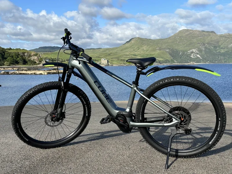 Electric mountain bike parked on a paved area near a body of water with mountains in the background. The bike is black and gray with green accents.