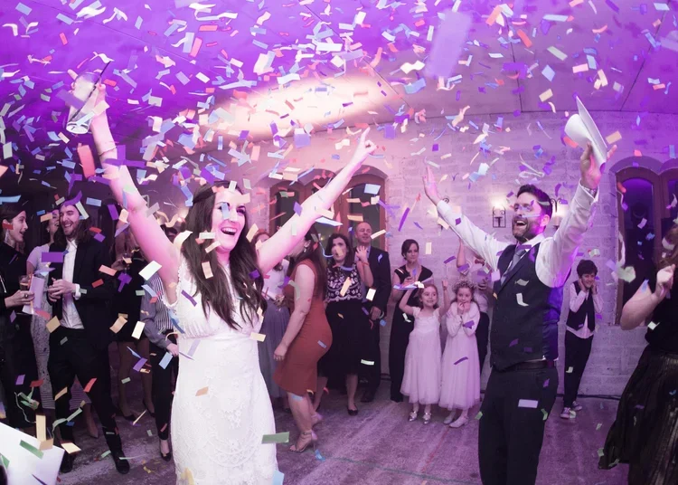 A celebration with people throwing colorful confetti in a room with stone walls, some dressed in formal attire, including a bride and groom dancing.