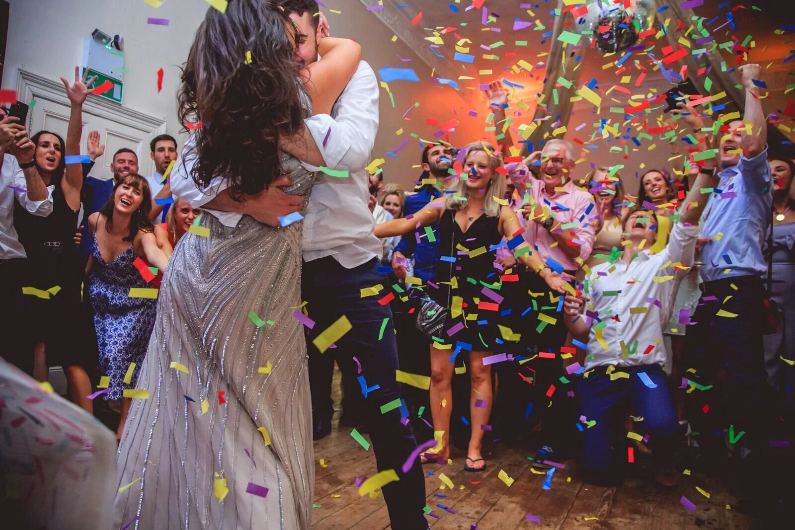 A wedding celebration with a bride and groom hugging in the center, surrounded by guests cheering and throwing colorful confetti in the air.