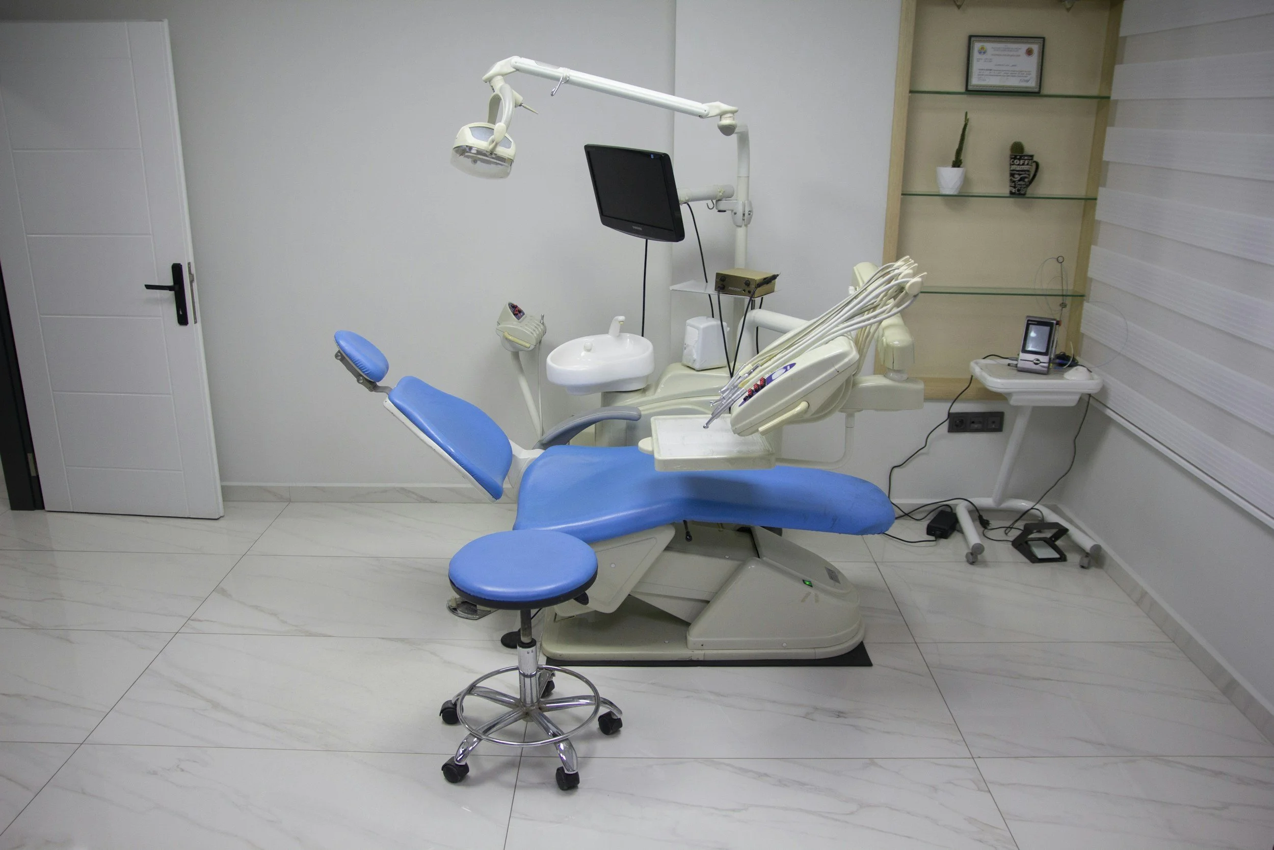 Dental examination chair in a clinic room with a stool, attached lights, monitor, and dental tools.