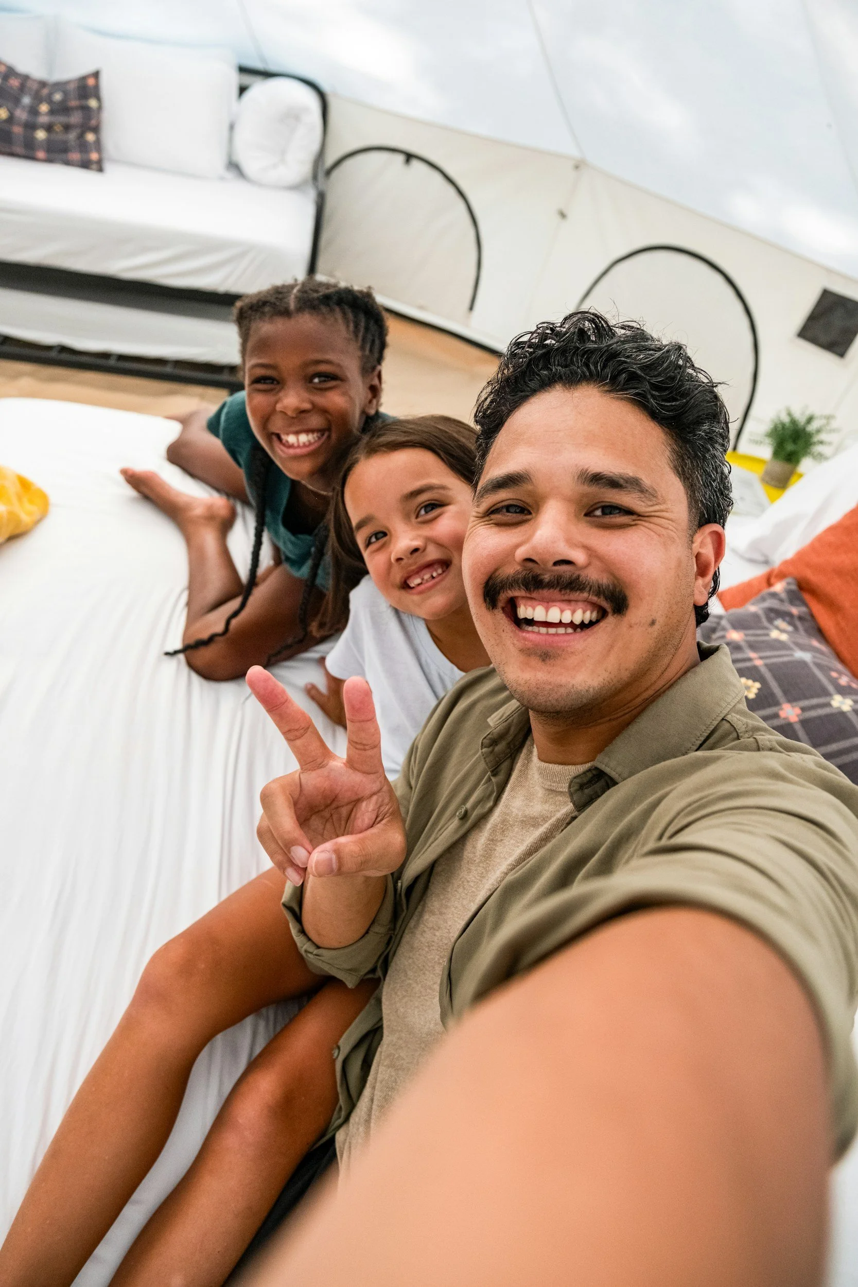 A man taking a selfie with two smiling children inside a tent, showing joyful expressions and peace sign gesture.