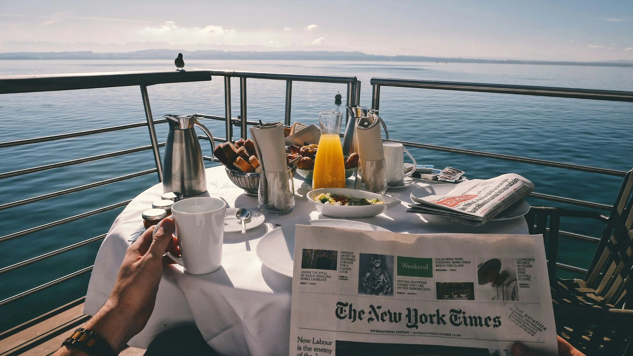 A dining setup on a boat deck with a table covered in a white tablecloth, featuring a breakfast spread including orange juice, coffee, bread, a salad, and a newspaper, overlooking calm water with a seagull perched on the railing and a distant shoreline under a partly cloudy sky.