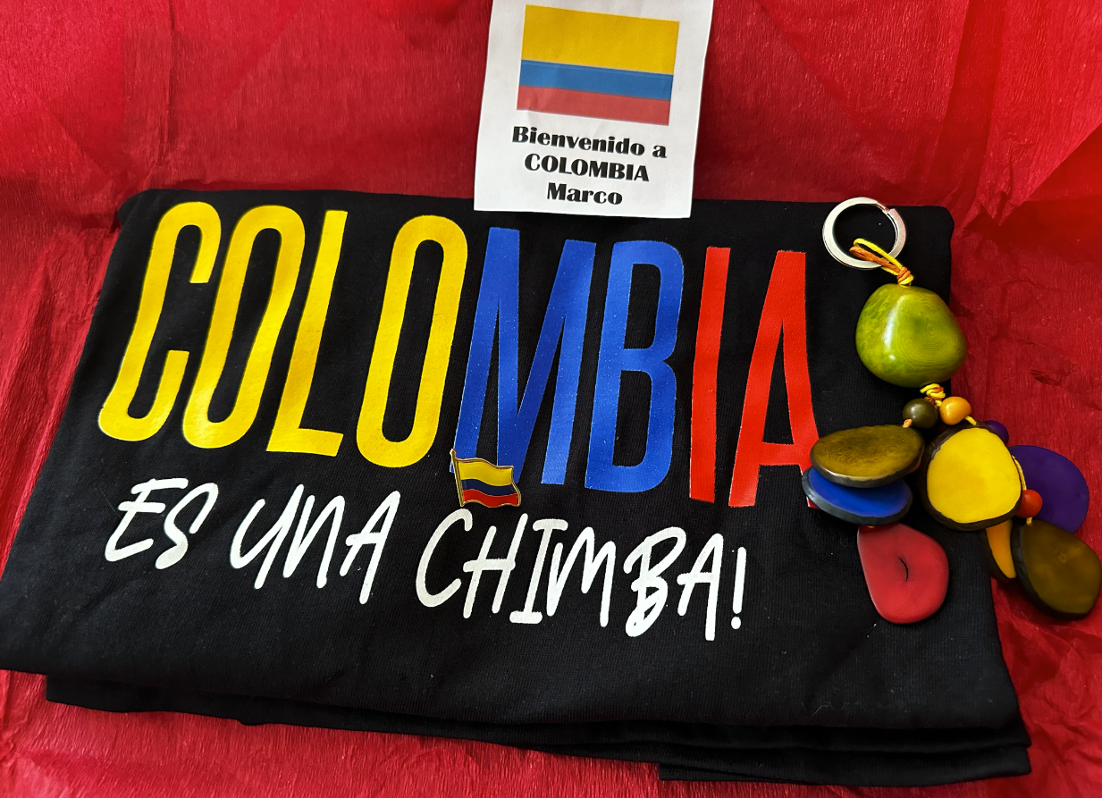 A black T-shirt with the words "COLOMBIA ES UNA CHIMBA" in colorful text, a small Colombian flag pin, and a keychain with yellow, green, blue, purple, and red beads, placed on a red surface with a Colombia welcome note.