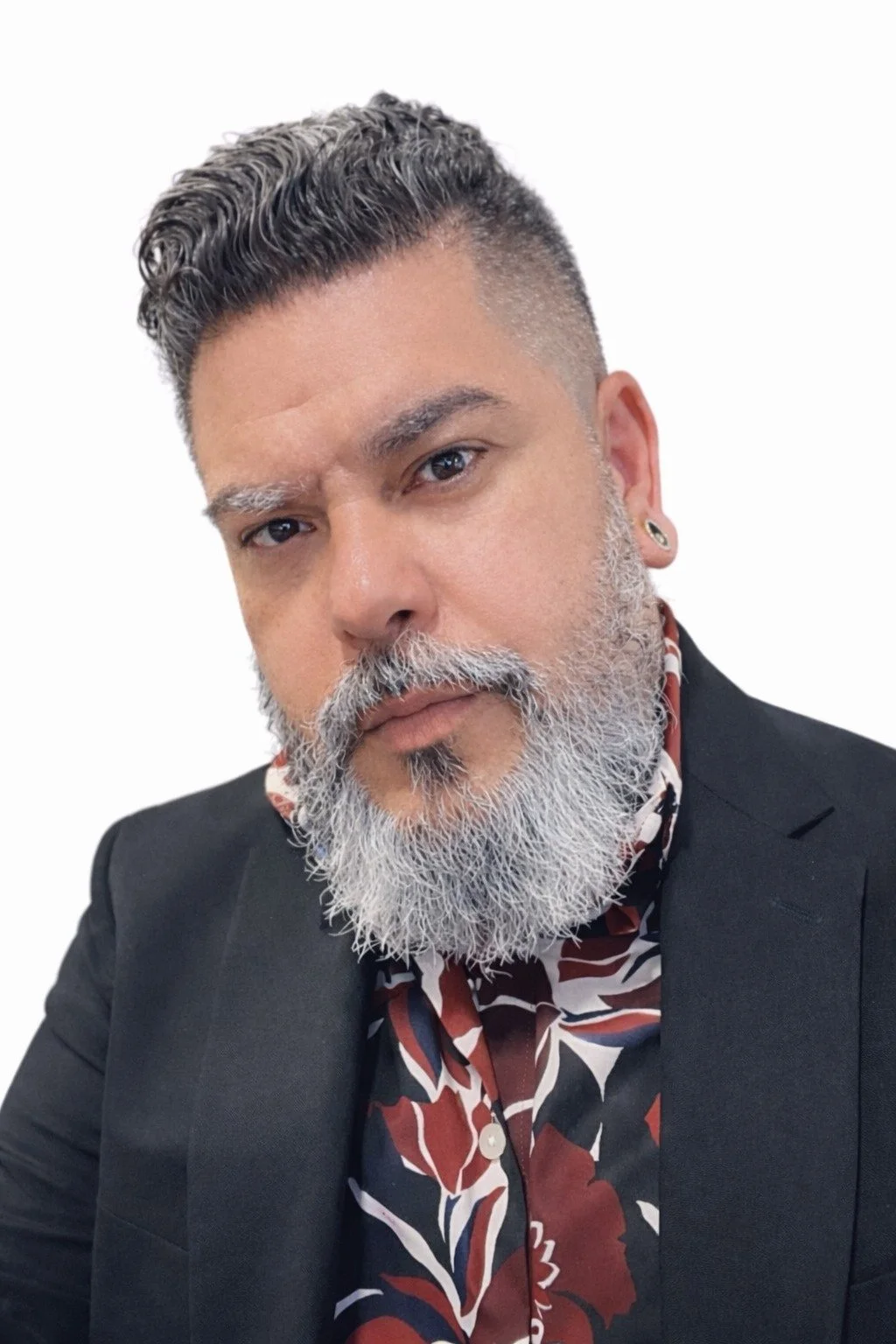 A man with a salt and pepper beard and styled hair wearing a black blazer over a patterned shirt, against a white background.