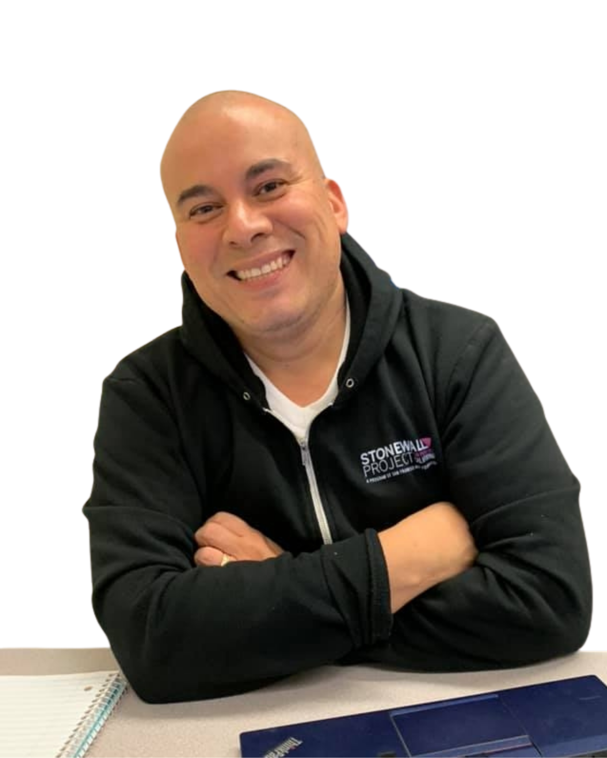 Smiling man with crossed arms, wearing a black hoodie with 'Stonewall Project' logo, sitting at a desk with a notebook and laptop.