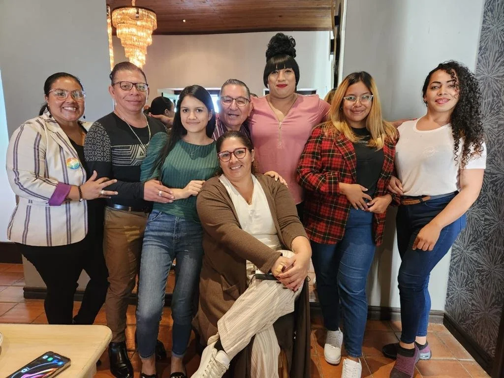 A group of nine women standing together indoors, smiling for the photo. Some are wearing glasses, and one is sitting in front with a brown jacket. The background shows a chandelier and a wall with patterned wallpaper.