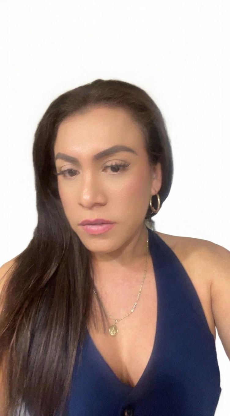 Portrait of a woman with long dark hair, wearing gold hoop earrings, a gold necklace, and a navy blue top against a white background.