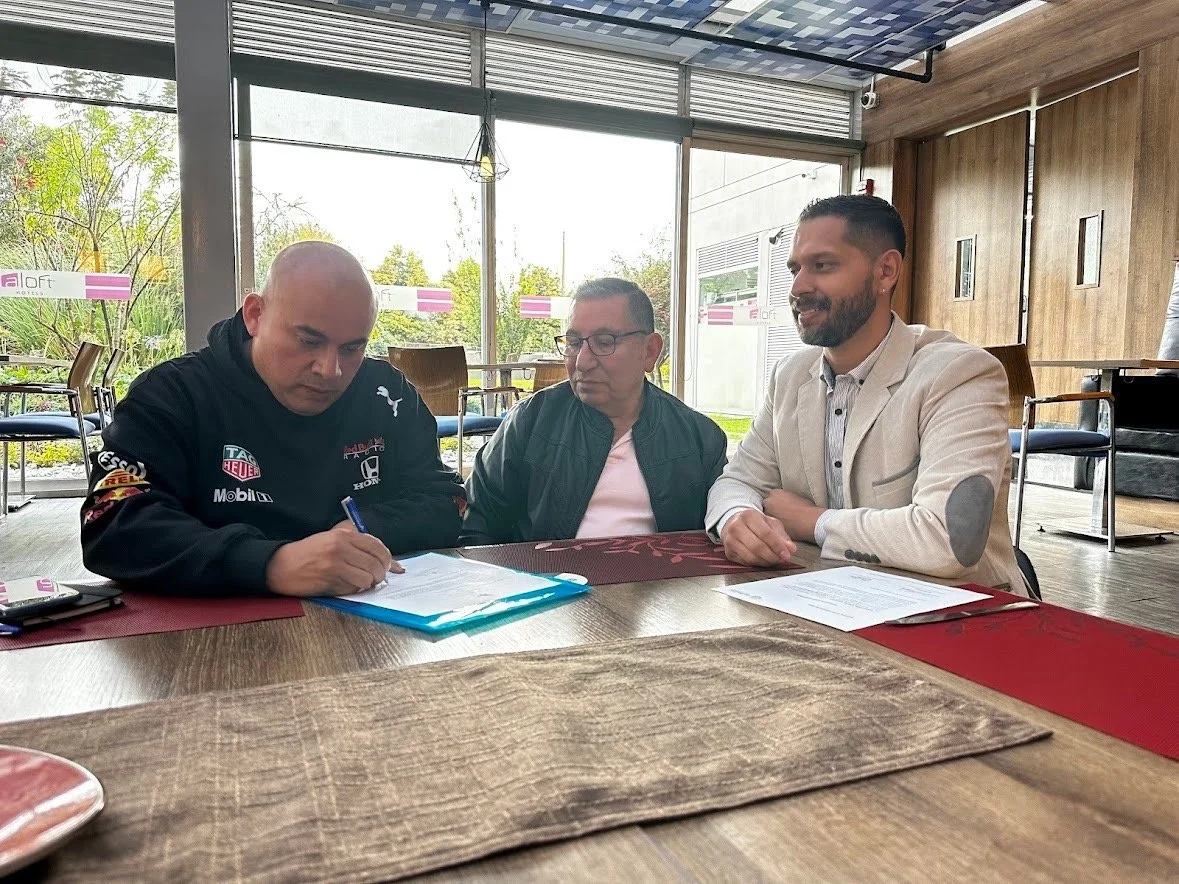 Three men sitting at a wooden table in a brightly lit restaurant or cafe, with the man on the left signing documents.