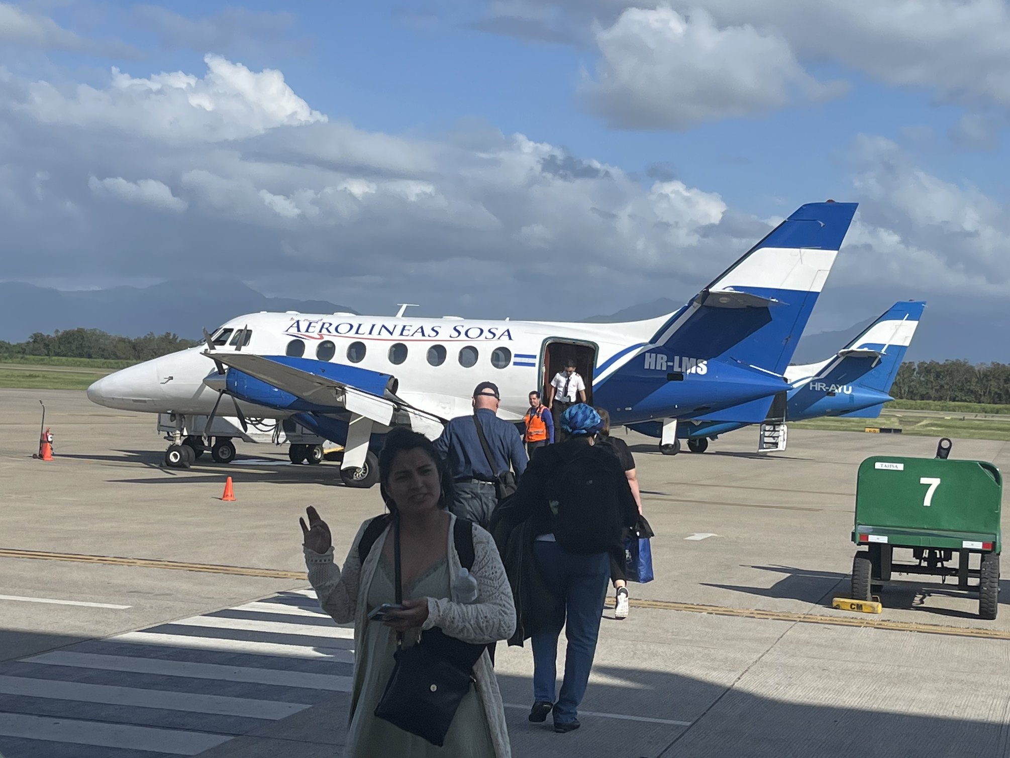 People boarding a small blue and white aircraft on an airport tarmac with a cloudy sky and mountains in the background.