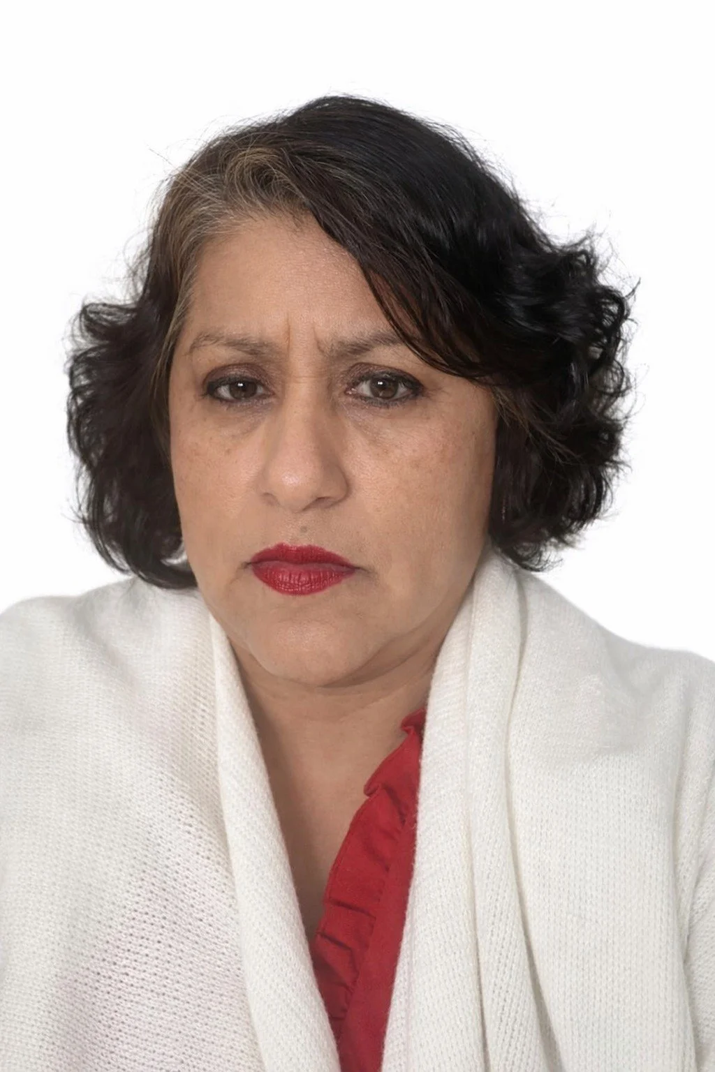 A middle-aged woman with dark, wavy hair and light skin, wearing a white blazer over a red top, looking directly at the camera with a neutral expression.