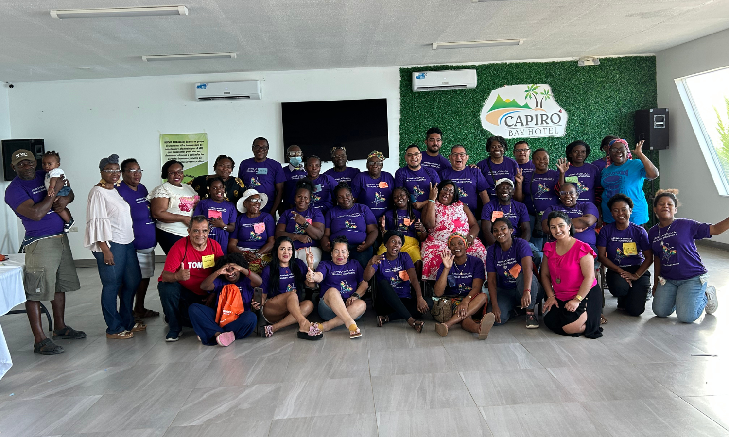 Group of people posing together indoors with a hotel sign in the background, some wearing matching purple T-shirts.