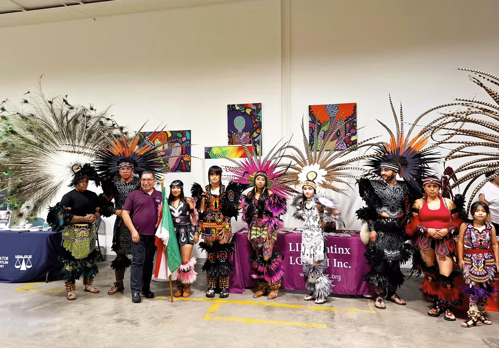 A group of Indigenous people in traditional attire and feathered headdresses standing in a line for a cultural event, with a person holding an Italian flag in the center.
