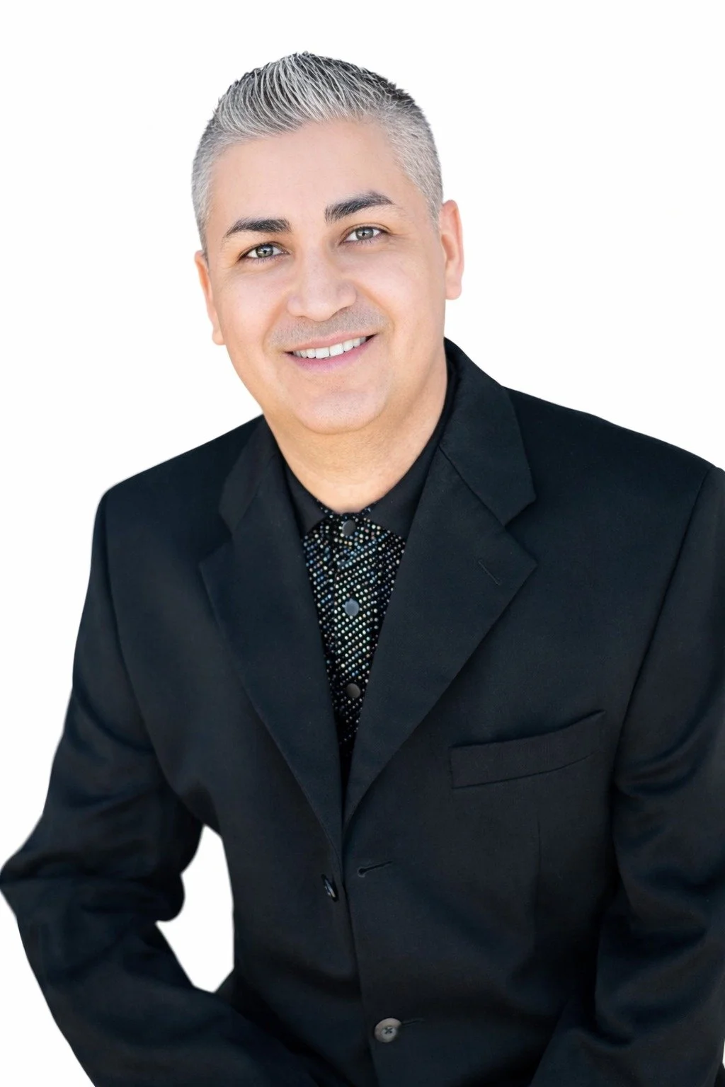 Portrait of a smiling man with short gray hair, wearing a black suit and a patterned black shirt, isolated on white background.