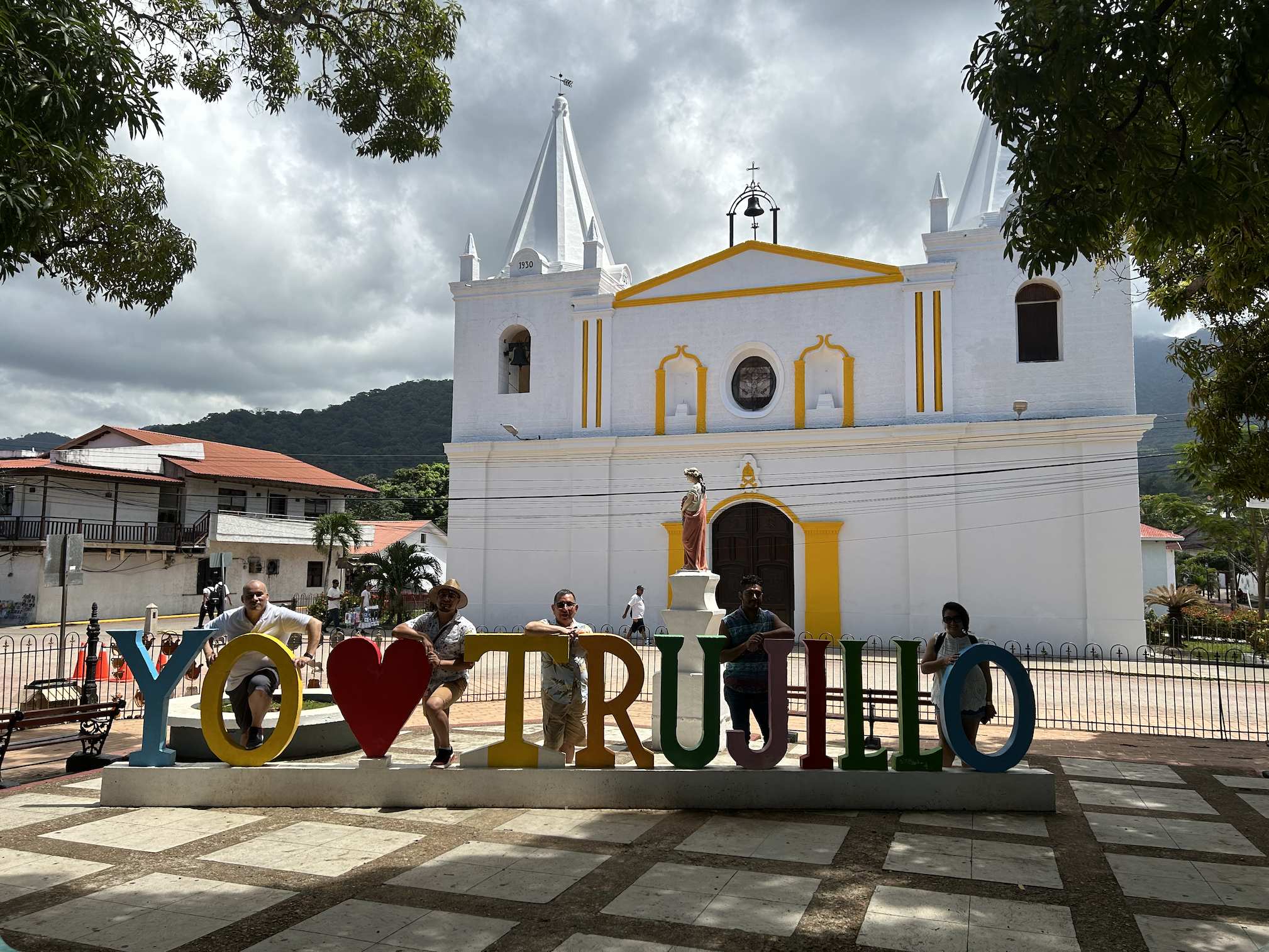 People posing in front of a church with colorful letters spelling 'Y O ❤️ R U S I L L O' on a bench.