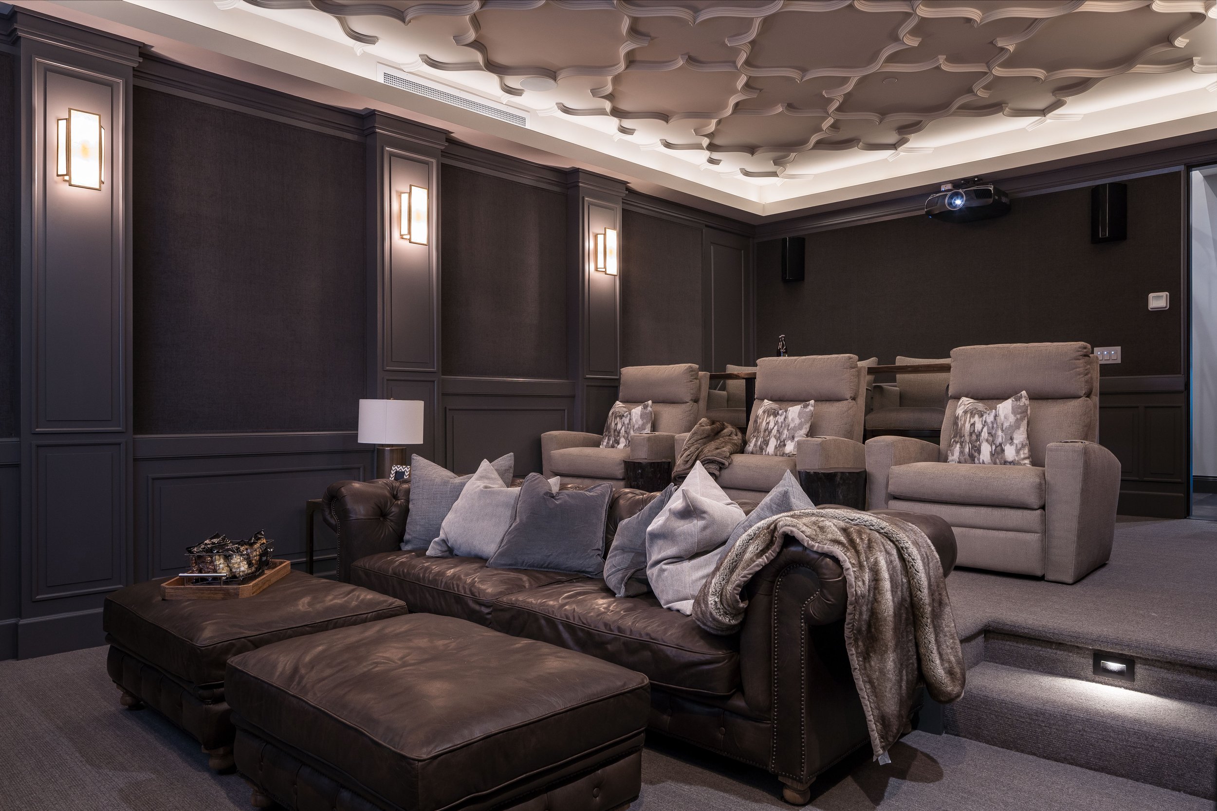 Home theater room with dark gray walls, ceiling with decorative panels, beige recliner chairs, gray sofa with pillows, side table, coffee table, and a projector mounted on the ceiling.