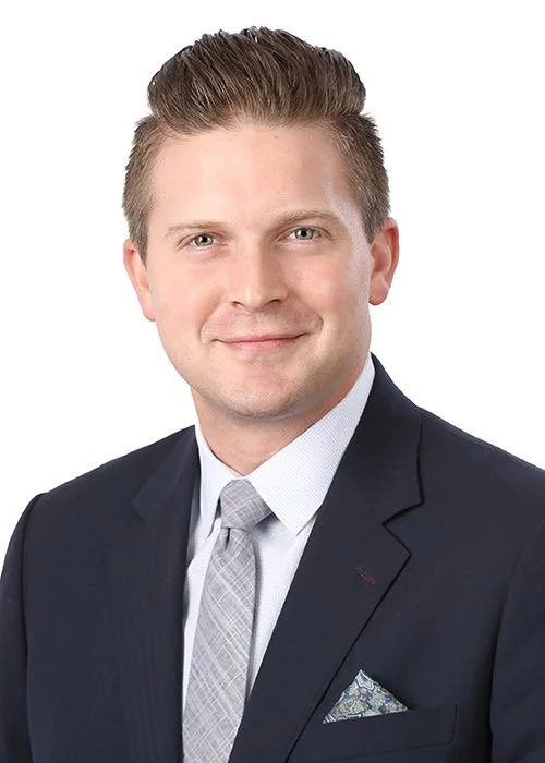Headshot of a young man in a dark suit, light shirt, and patterned tie, smiling against a white background.