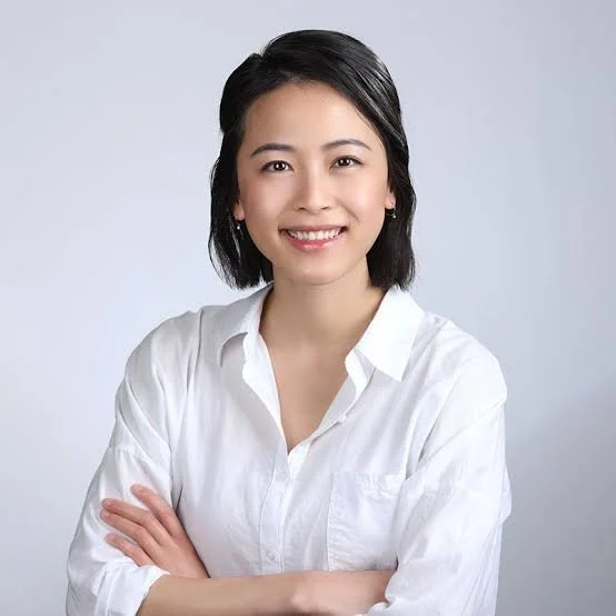 A young woman with short black hair, smiling, wearing a white button-up shirt, crossing her arms, against a light gray background.